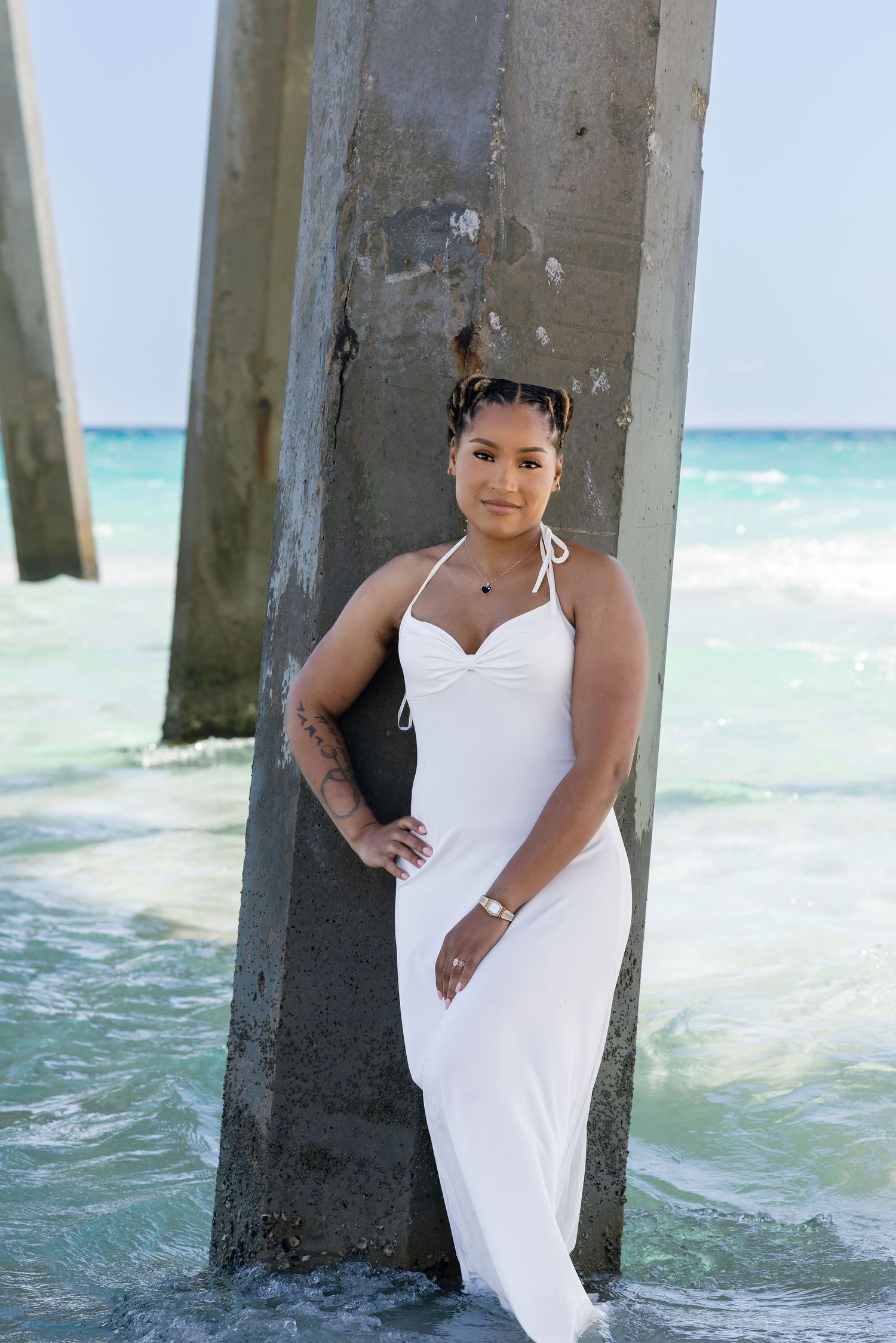 Woman in white maxi dress poses by beach pier pillars with turquoise ocean waves in background.