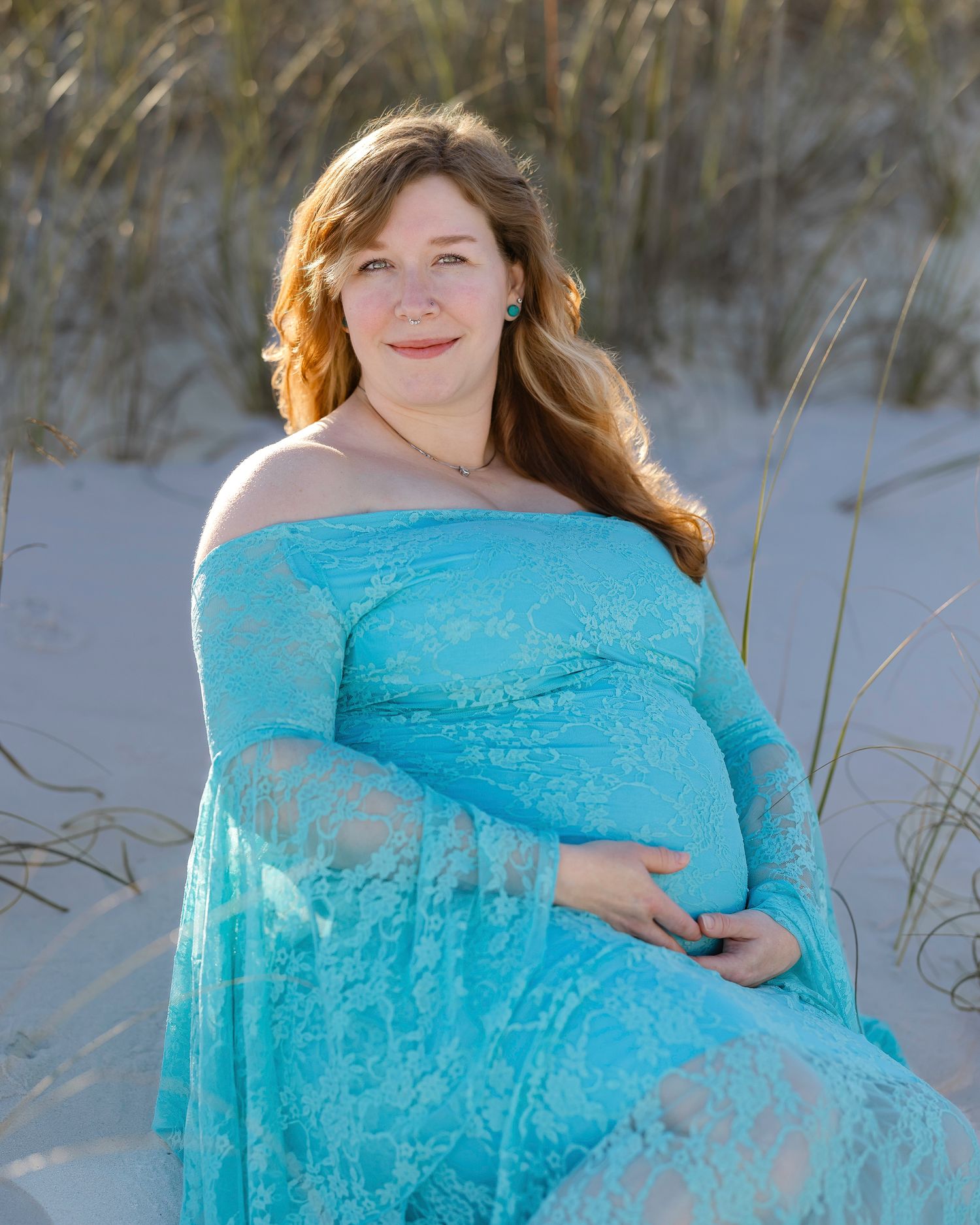 Pregnant woman in turquoise lace off-shoulder gown sitting on sandy beach dunes, cradling baby bump.