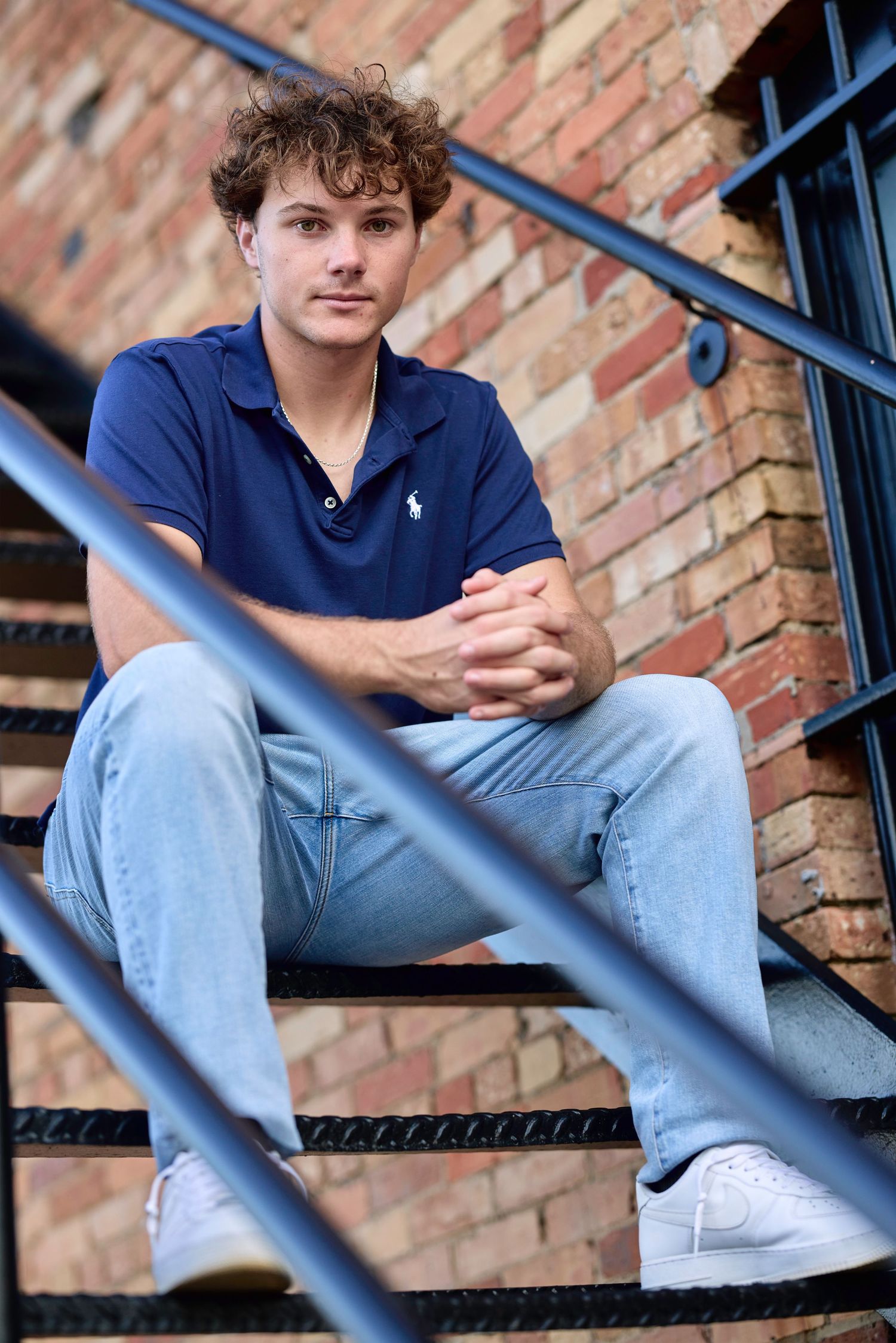 Young man in navy polo and jeans sits on metal stairs against brick wall.