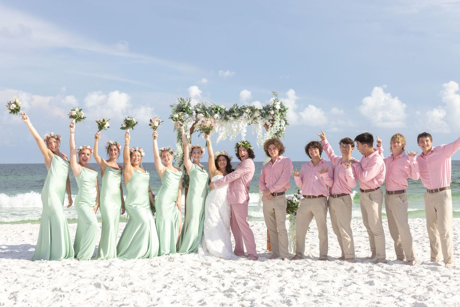 Wedding party in pastel attire lined up on beach under blue sky.