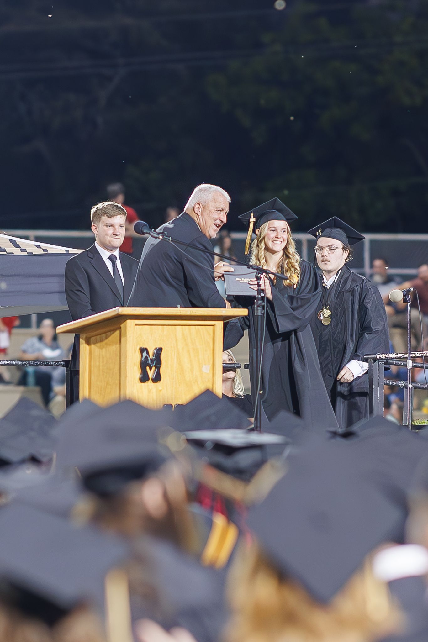 A graduation ceremony speaker addresses the audience from a wooden podium during an outdoor commencement.
