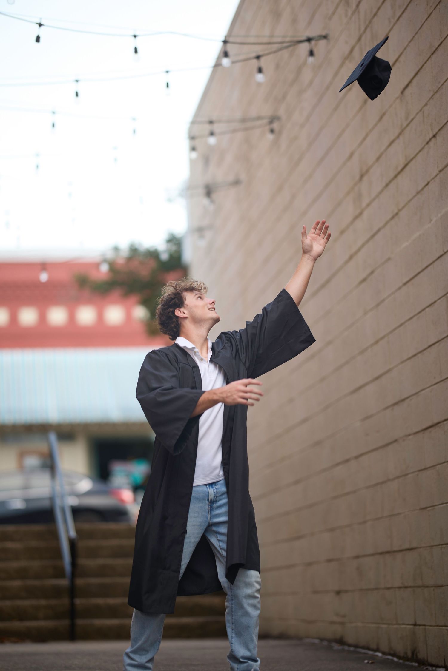 Graduate in black gown tossing cap in celebration against brick wall with string lights.