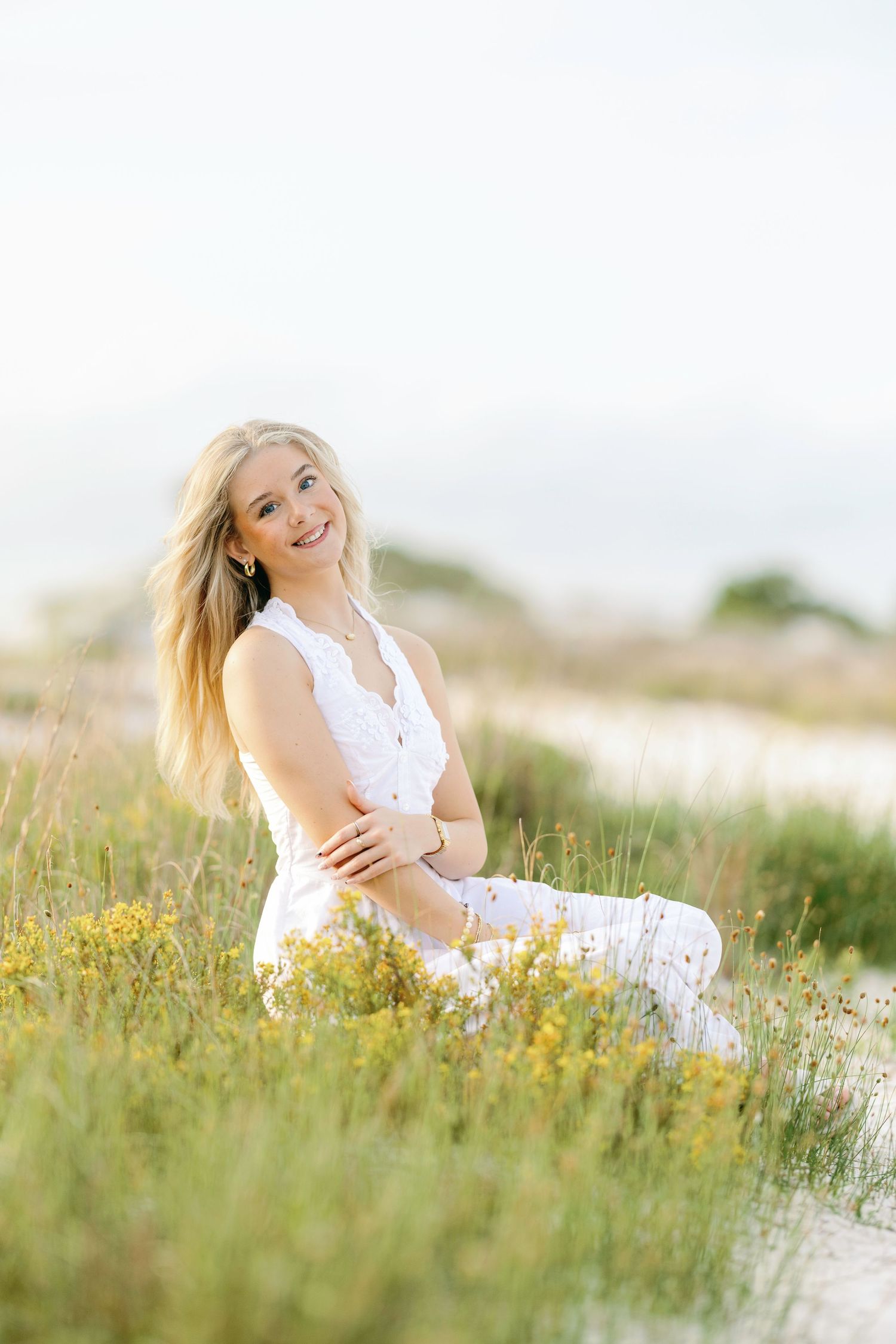 Graduate in white dress sitting among coastal wildflowers and dunes.