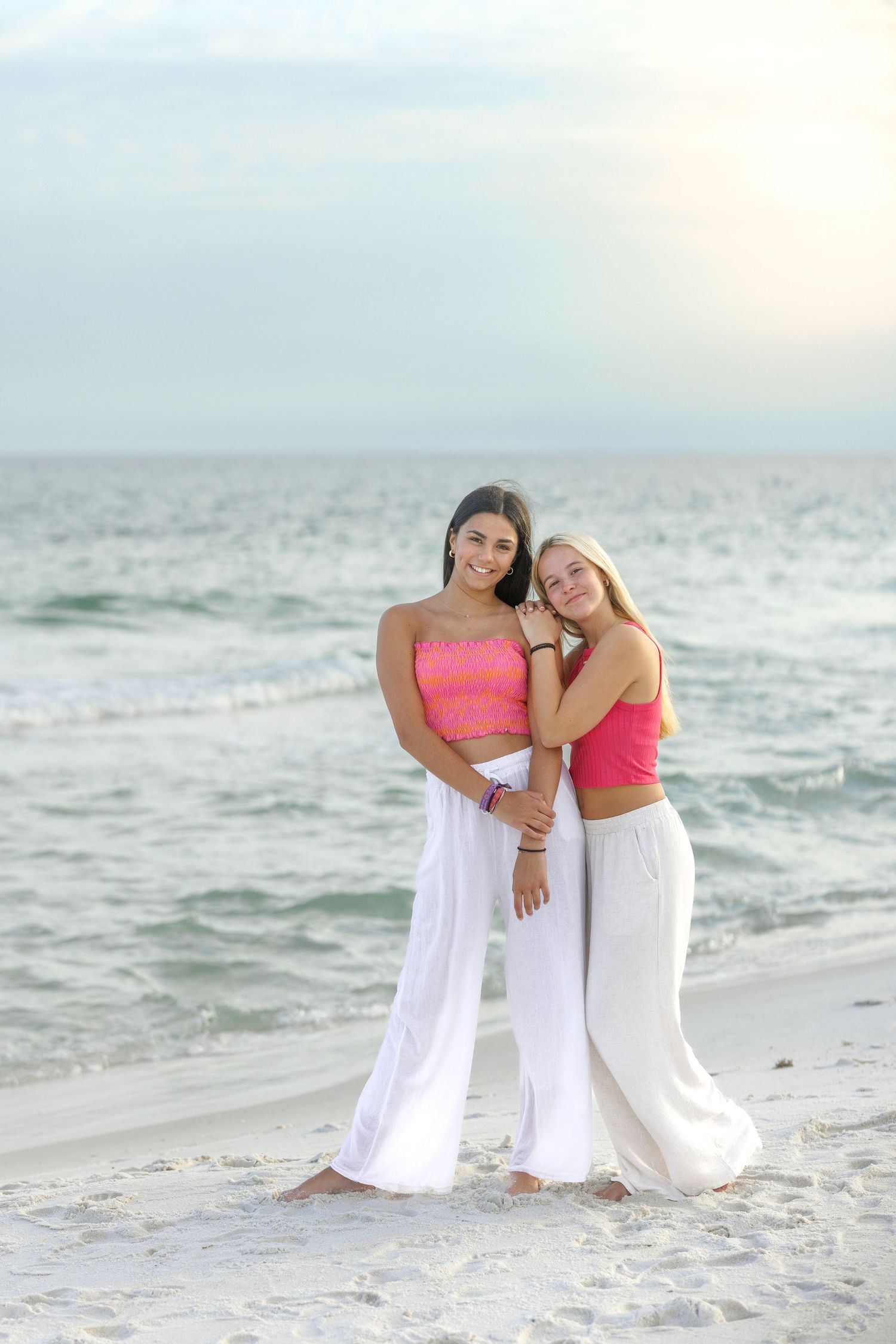Friends in coordinated beachwear embracing on white sand with ocean waves behind them.