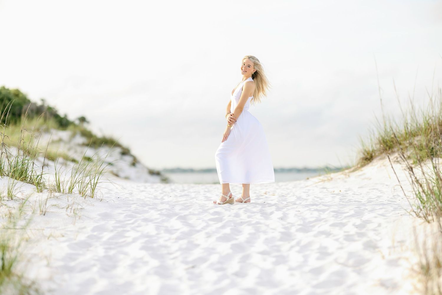 Beachside photoshoot featuring a flowing white dress against the ocean waves and sand dunes at sunset.