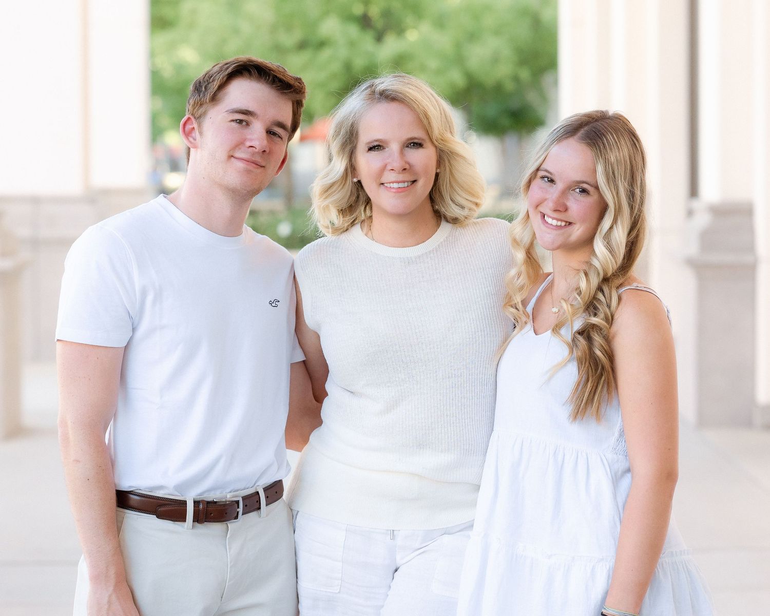 Three smiling people in white outfits pose together outdoors near columns.