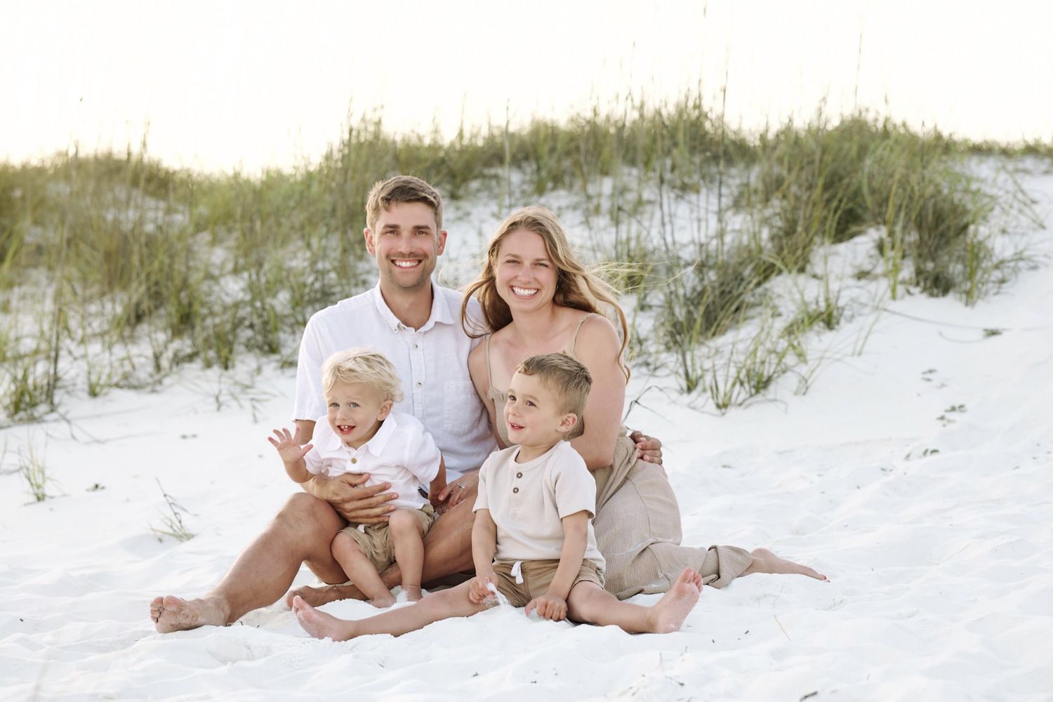 Happy family of four posing together on white sand beach dunes, parents with two young blonde boys smiling.