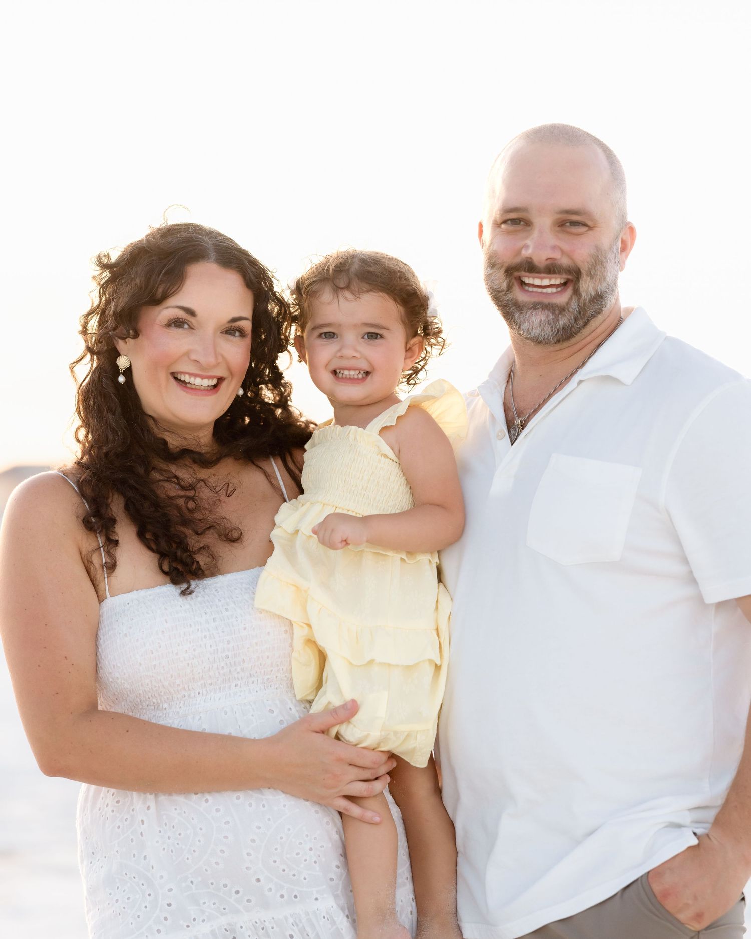 Happy family of three posing on bright sunny beach, parents holding young daughter in yellow dress.