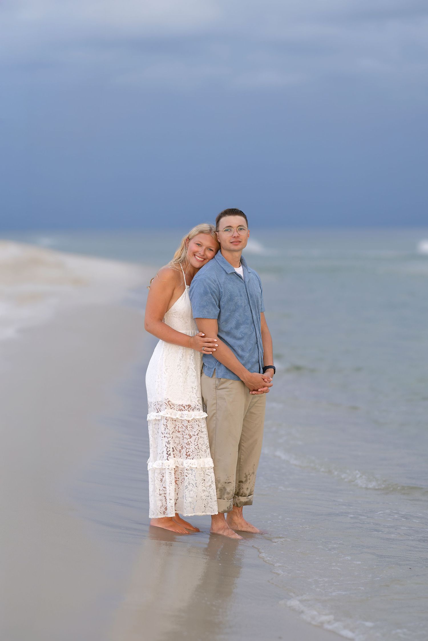 A smiling couple embraces on a sandy beach, woman in white dress and man in blue shirt, with ocean waves behind them.