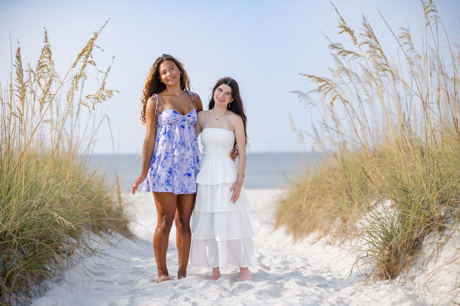 Two young women in summer dresses pose together on a sandy beach path framed by tall sea oats.
