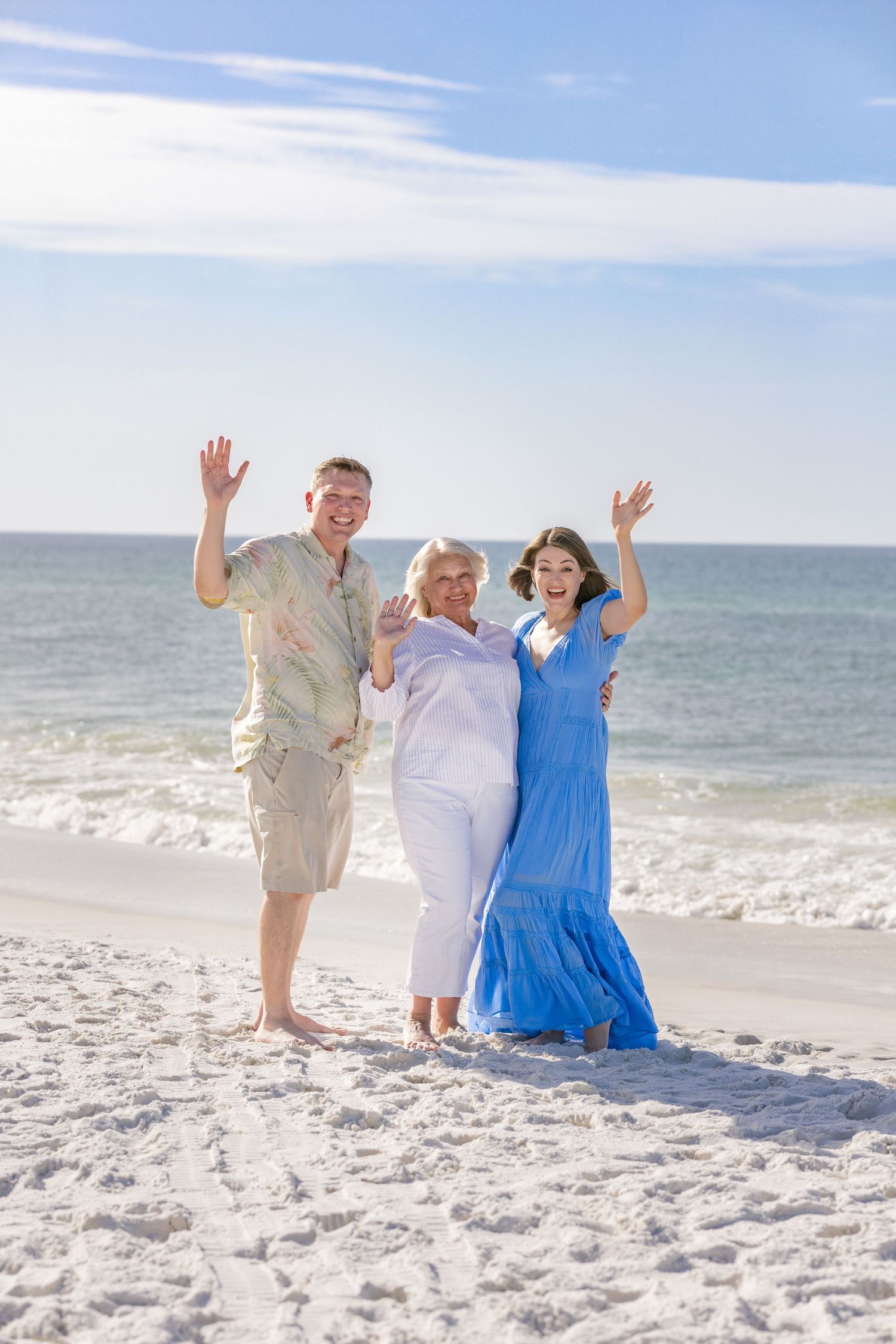 Three happy people waving on a sunny beach with ocean waves, wearing casual summer clothes on white sand.