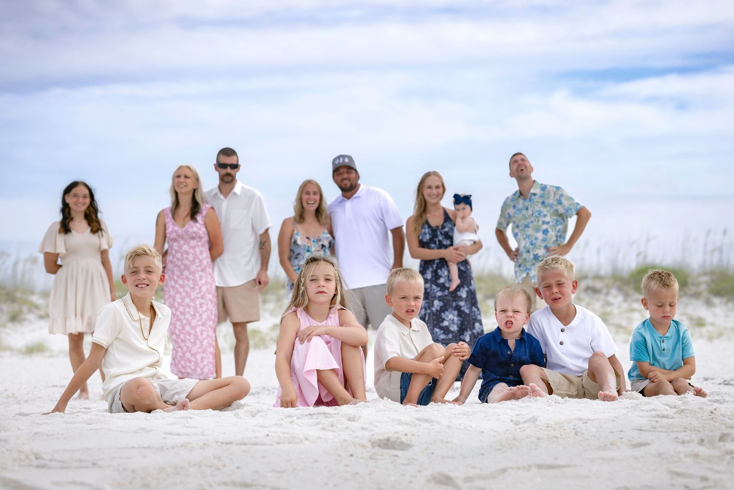 Large family group photo on white sand beach with adults standing and children sitting in front on a sunny day.