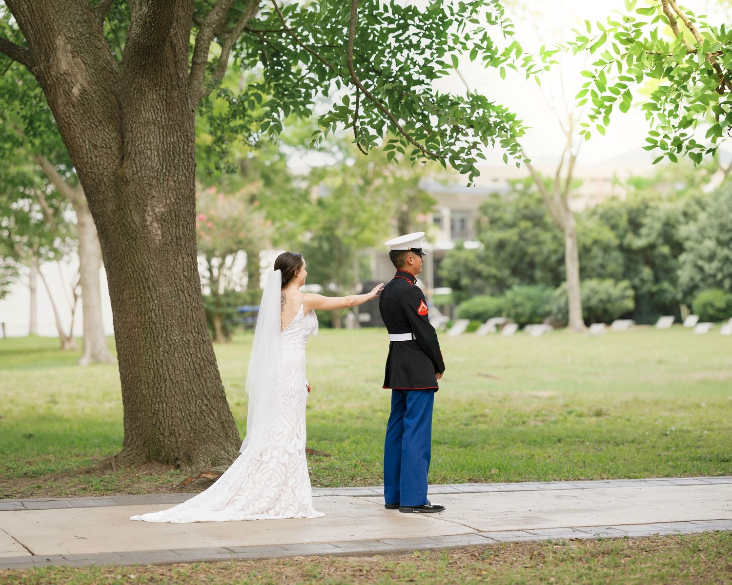 Military groom in dress uniform holds bride's hand under a large tree during outdoor wedding ceremony.