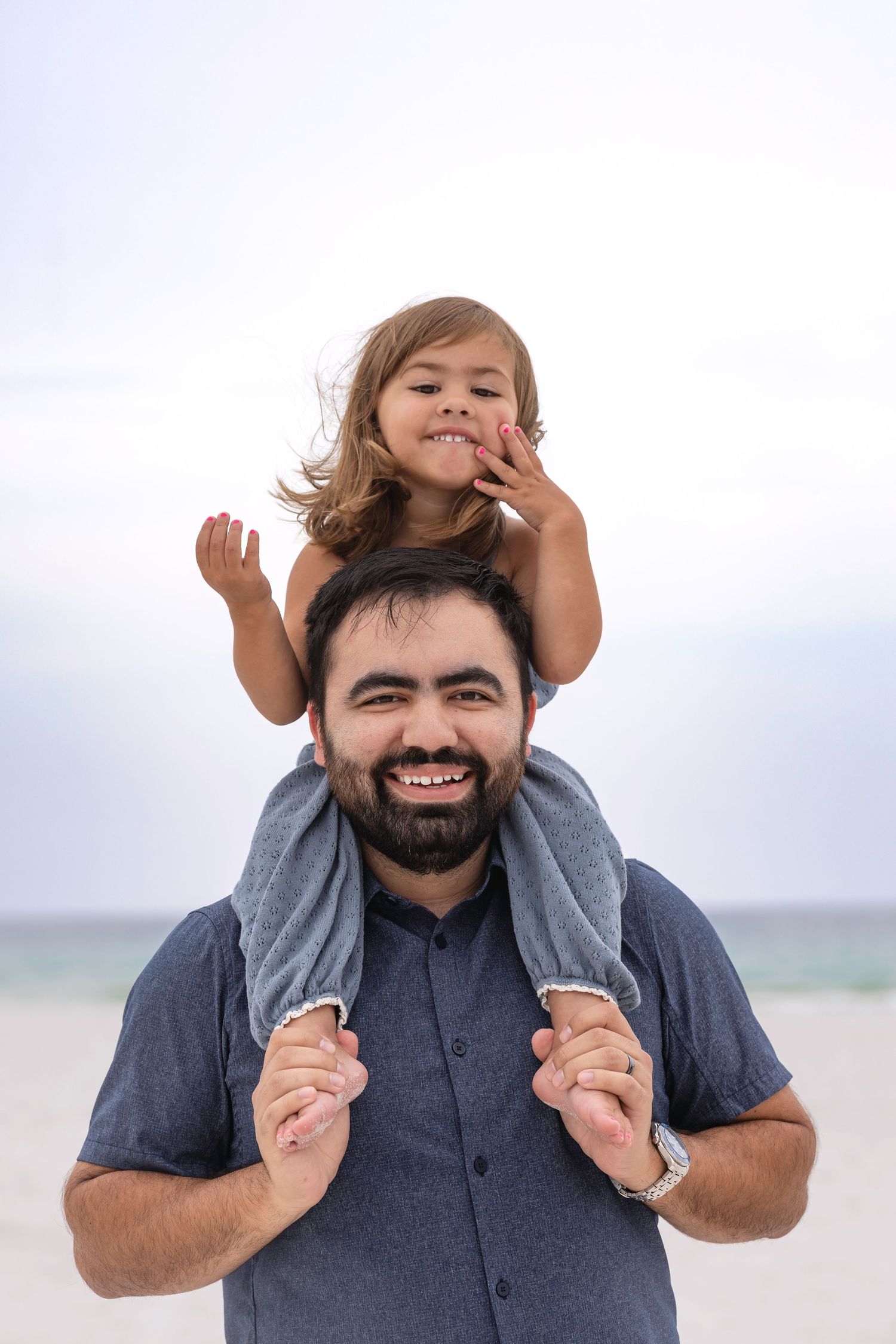 Smiling bearded man at beach carries happy young girl on his shoulders, both laughing outdoors.