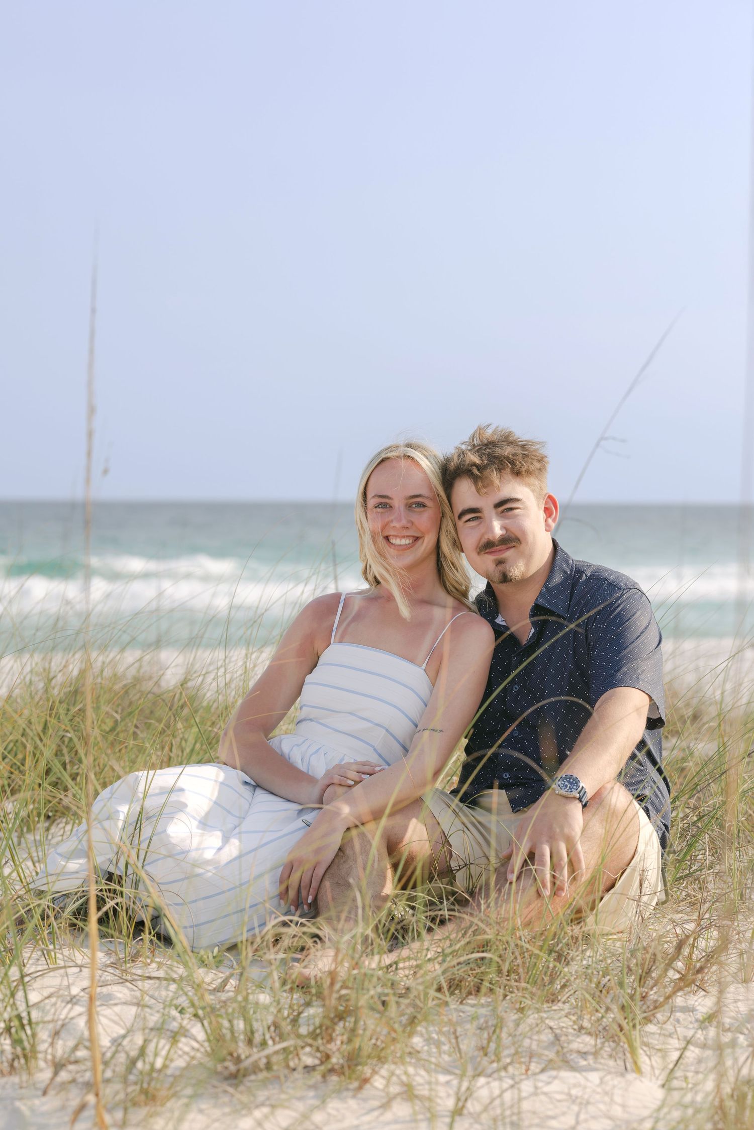 Smiling couple sitting together on sandy beach dunes with ocean waves in the background.