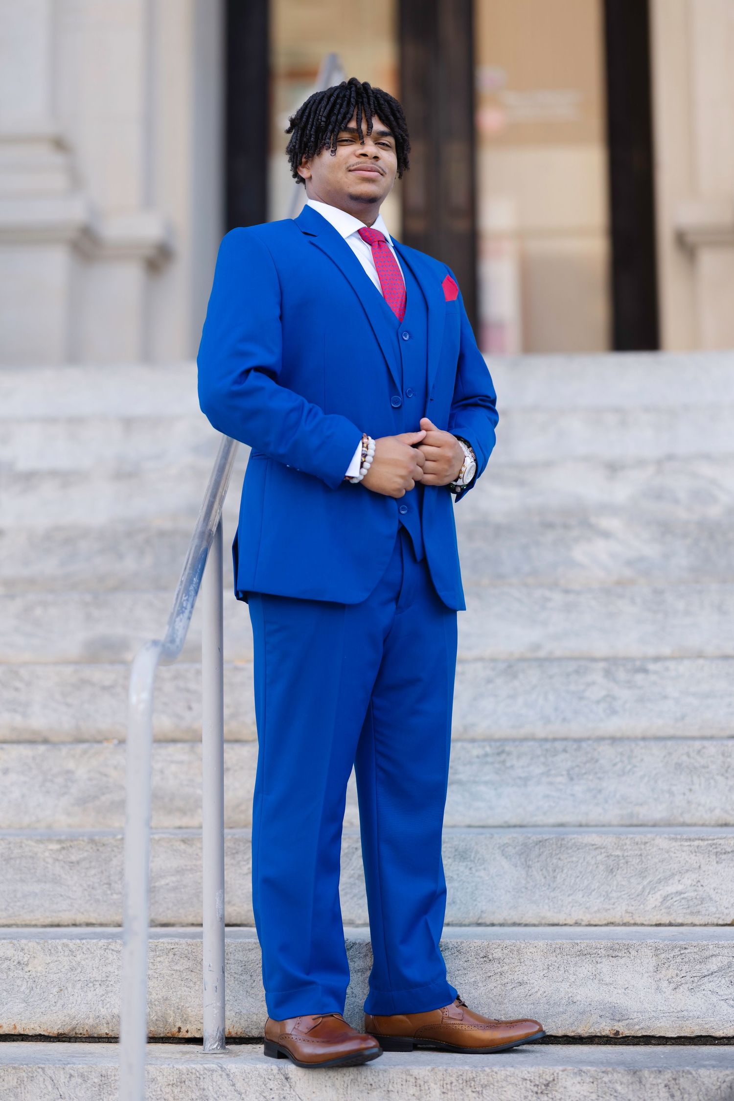 Young man in a bold royal blue three-piece suit with red tie and pocket square, posing on marble steps.