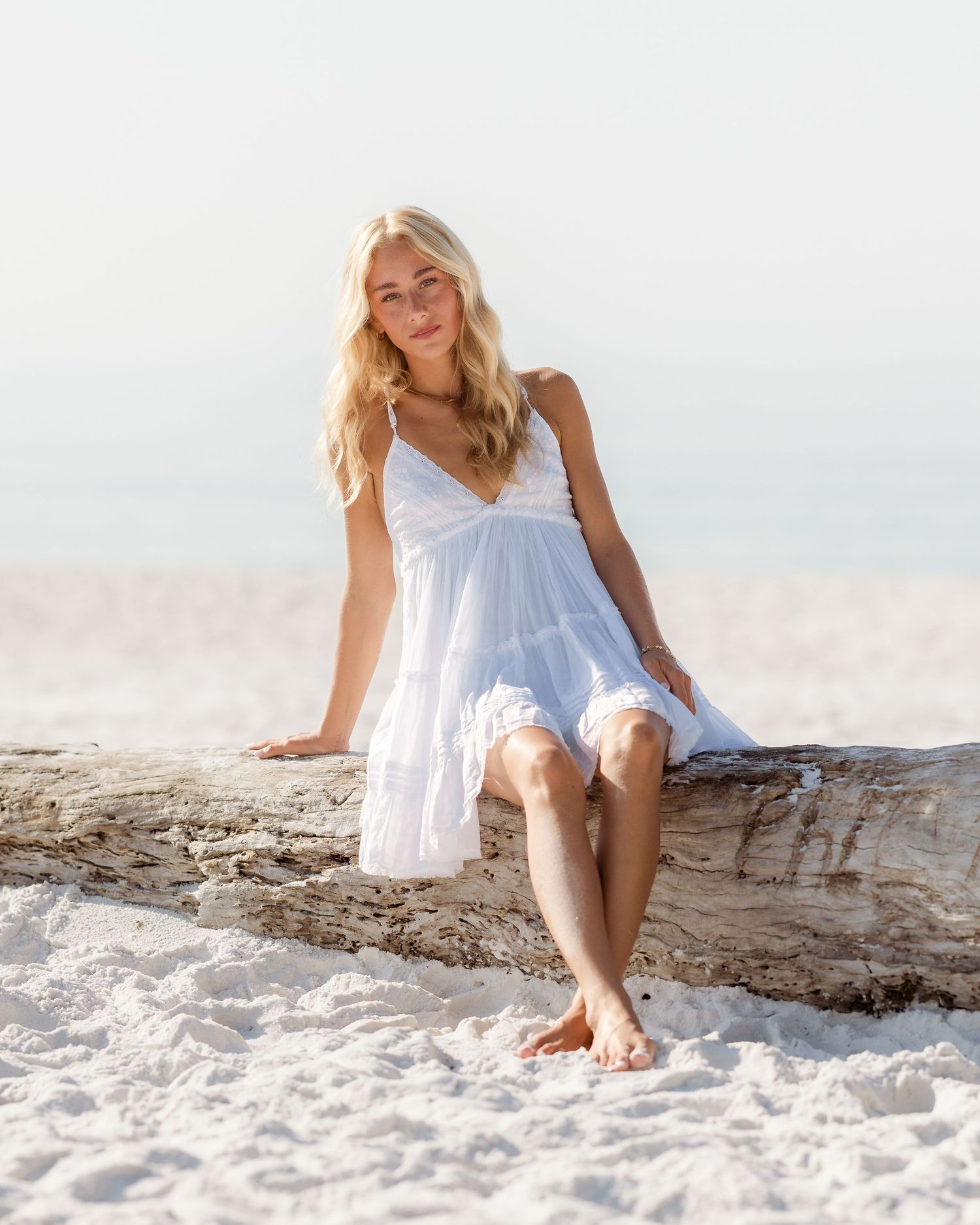 Blonde woman in white sundress sitting on driftwood on a white sand beach.