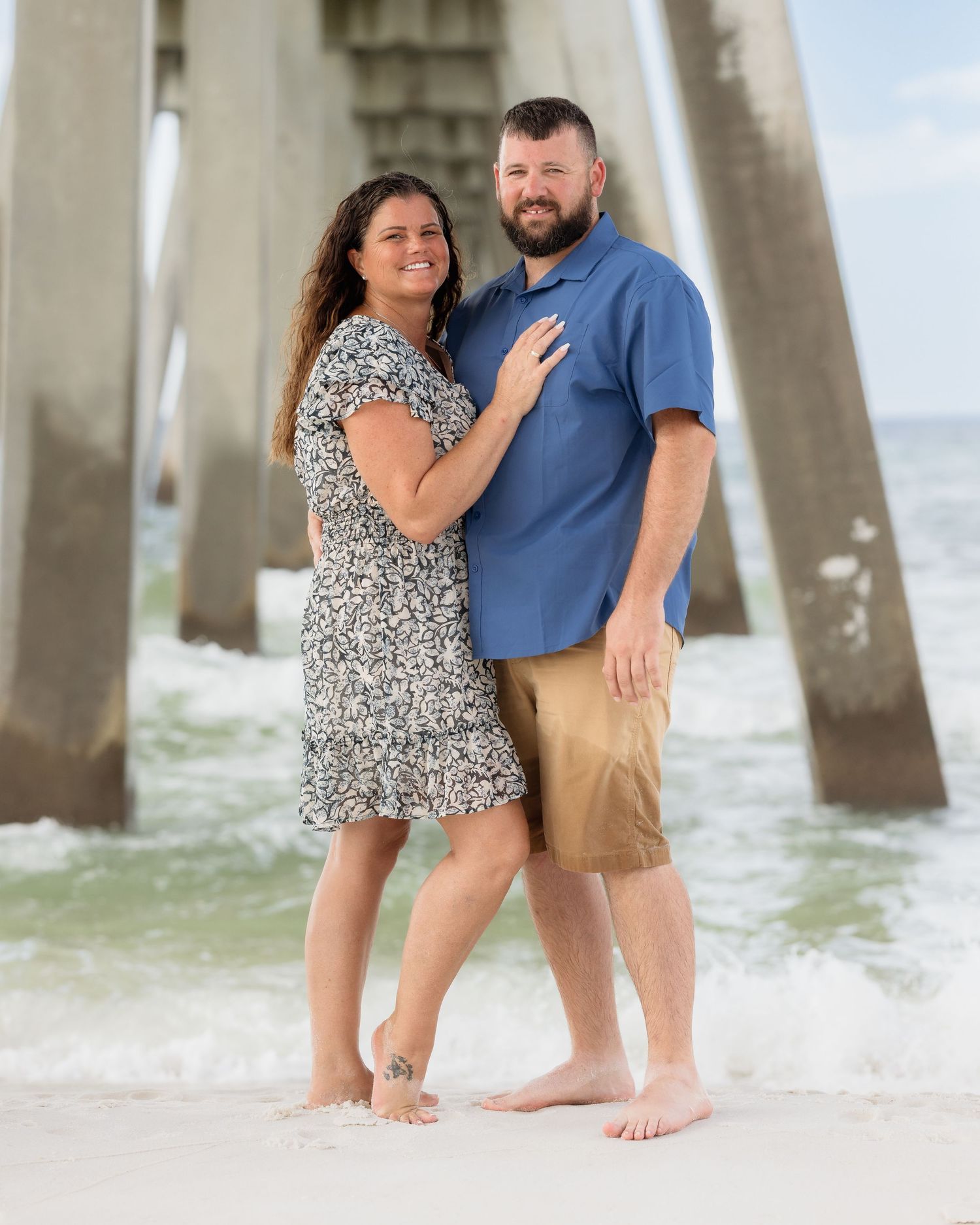 A smiling couple poses barefoot on the beach under a pier with ocean waves in the background.