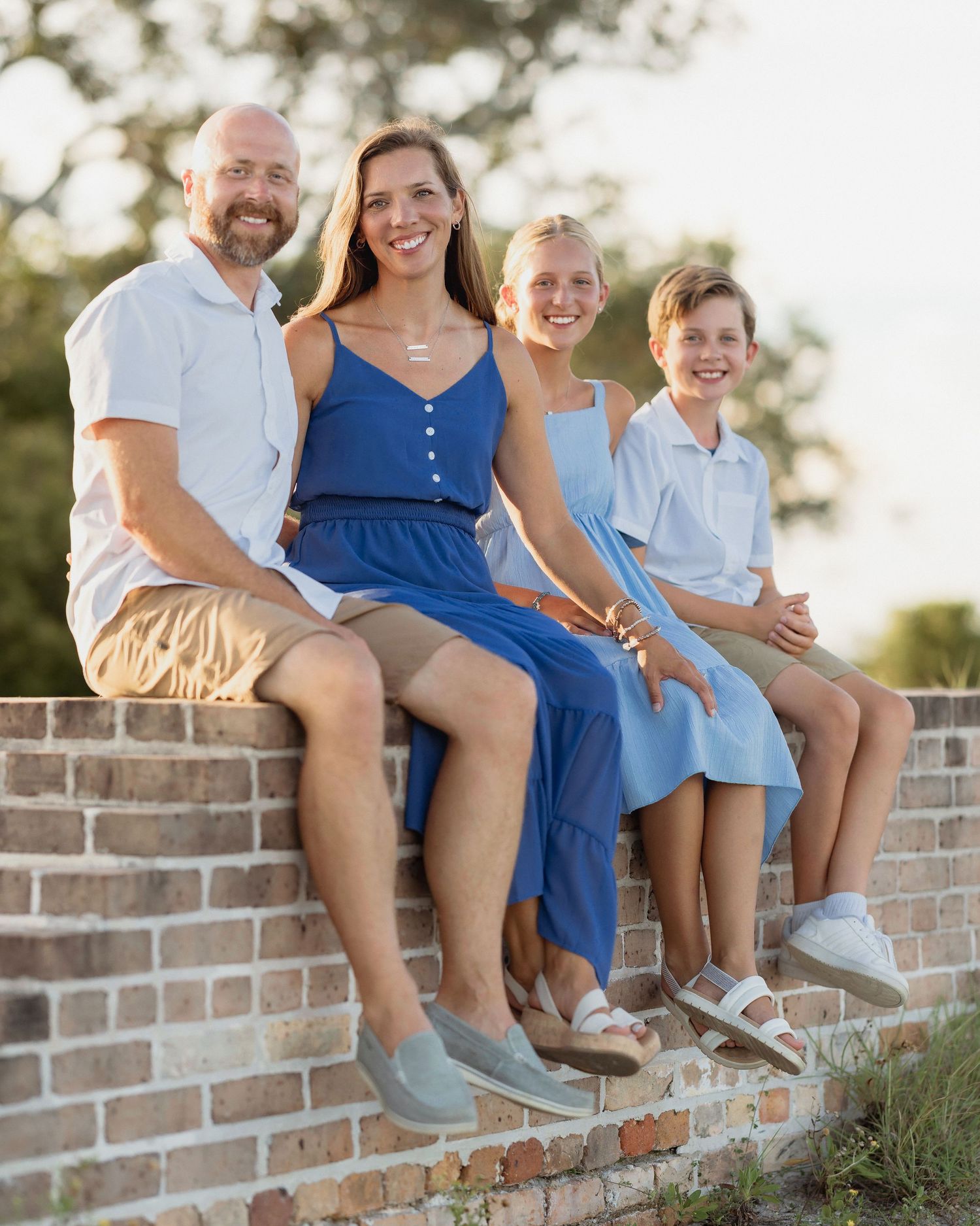 Family of four smiling and sitting on a brick wall outdoors during golden hour, wearing blue and white outfits.