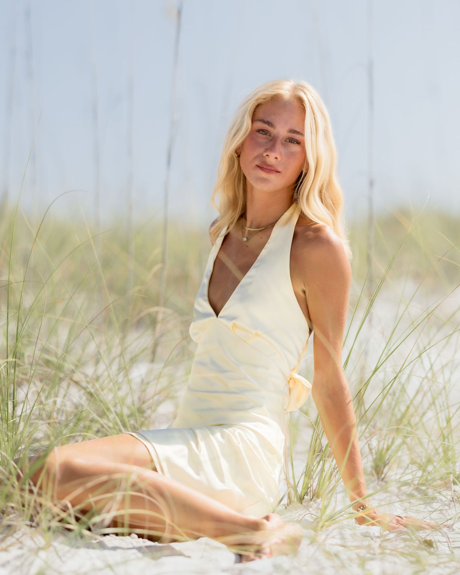 Blonde woman in yellow wrap dress sitting among sea oats on white sand dunes.