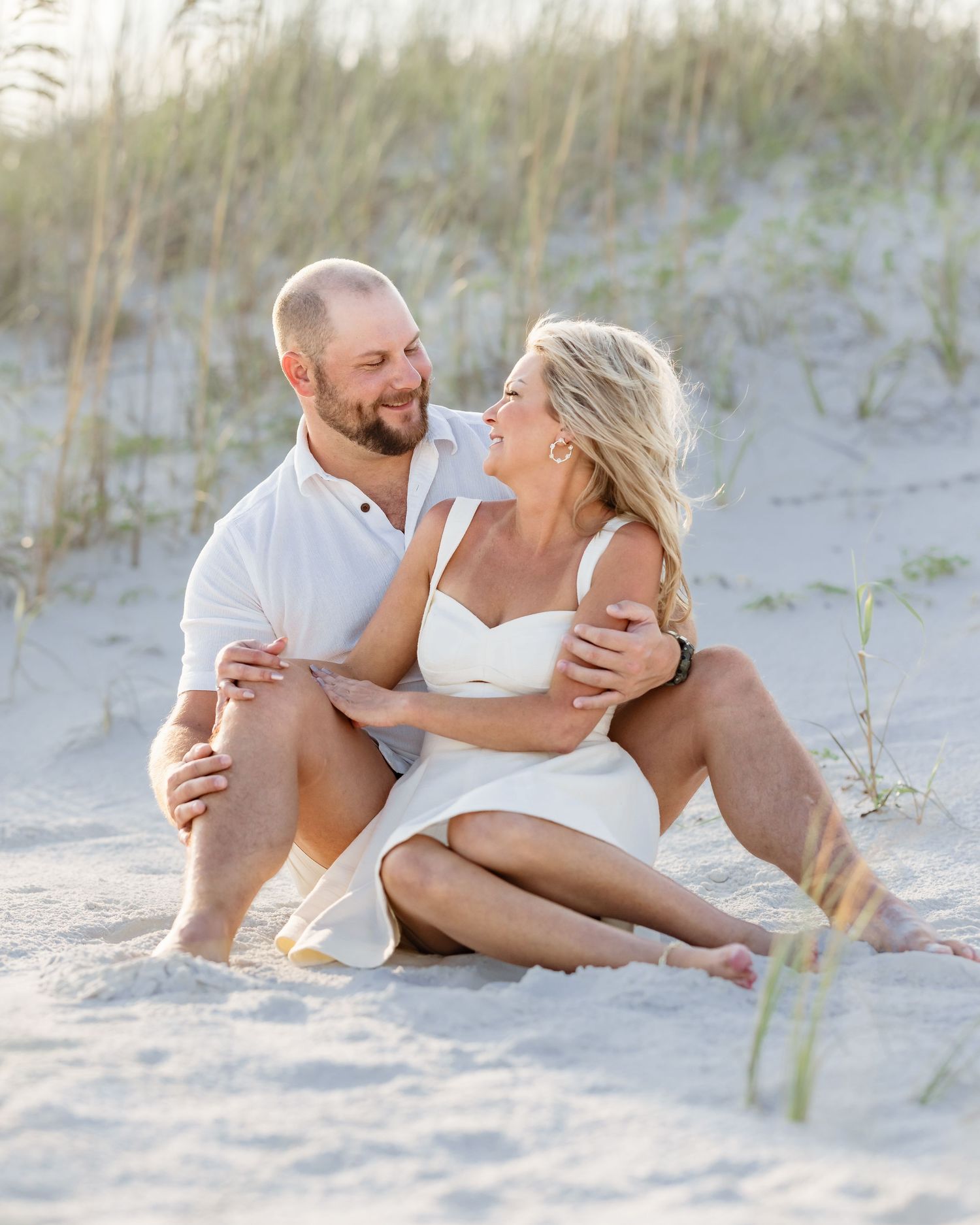 A couple sits together on sandy beach dunes, dressed in white, smiling at each other in a romantic engagement photo.