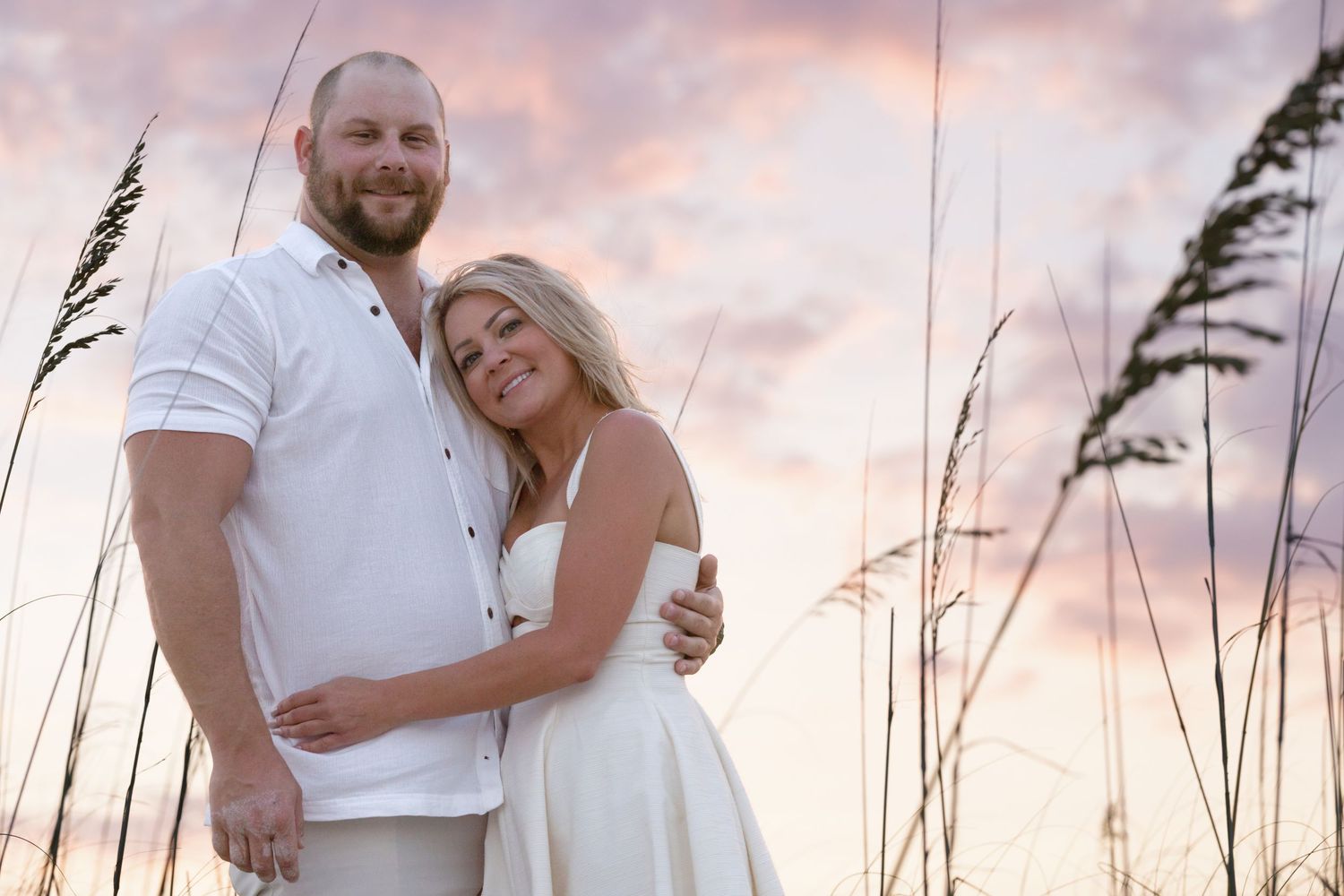 Couple embracing at sunset on a beach, wearing white outfits with sea oats and pastel sky behind them.