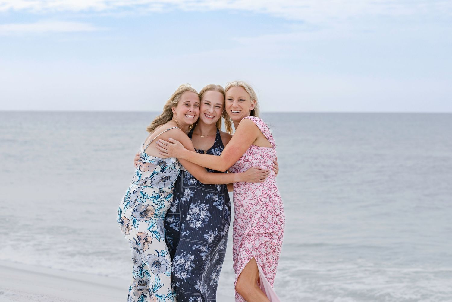 Three blonde women in floral dresses hugging and smiling on a beach with ocean waves in the background.