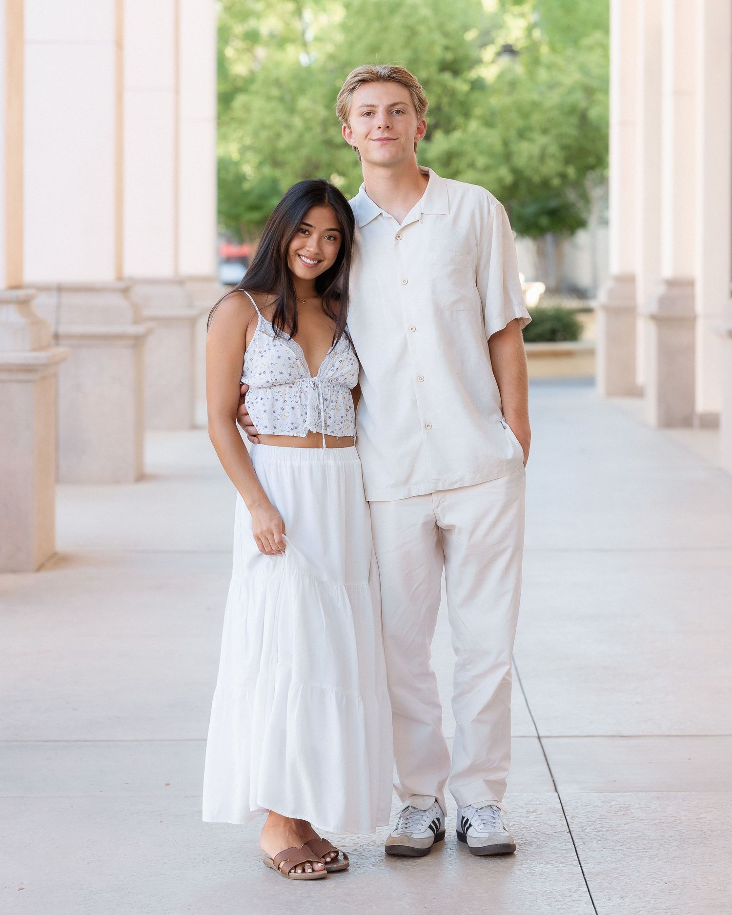 A young couple in matching white outfits posing together under an outdoor colonnade on a sunny day.