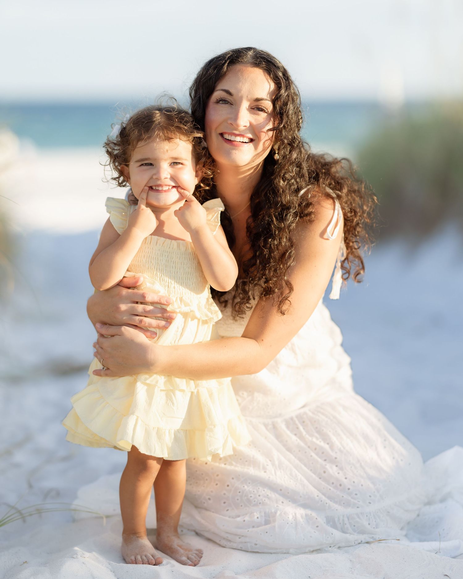 A smiling mother with curly dark hair holds her young daughter on a sandy beach, both wearing white and yellow dresses.