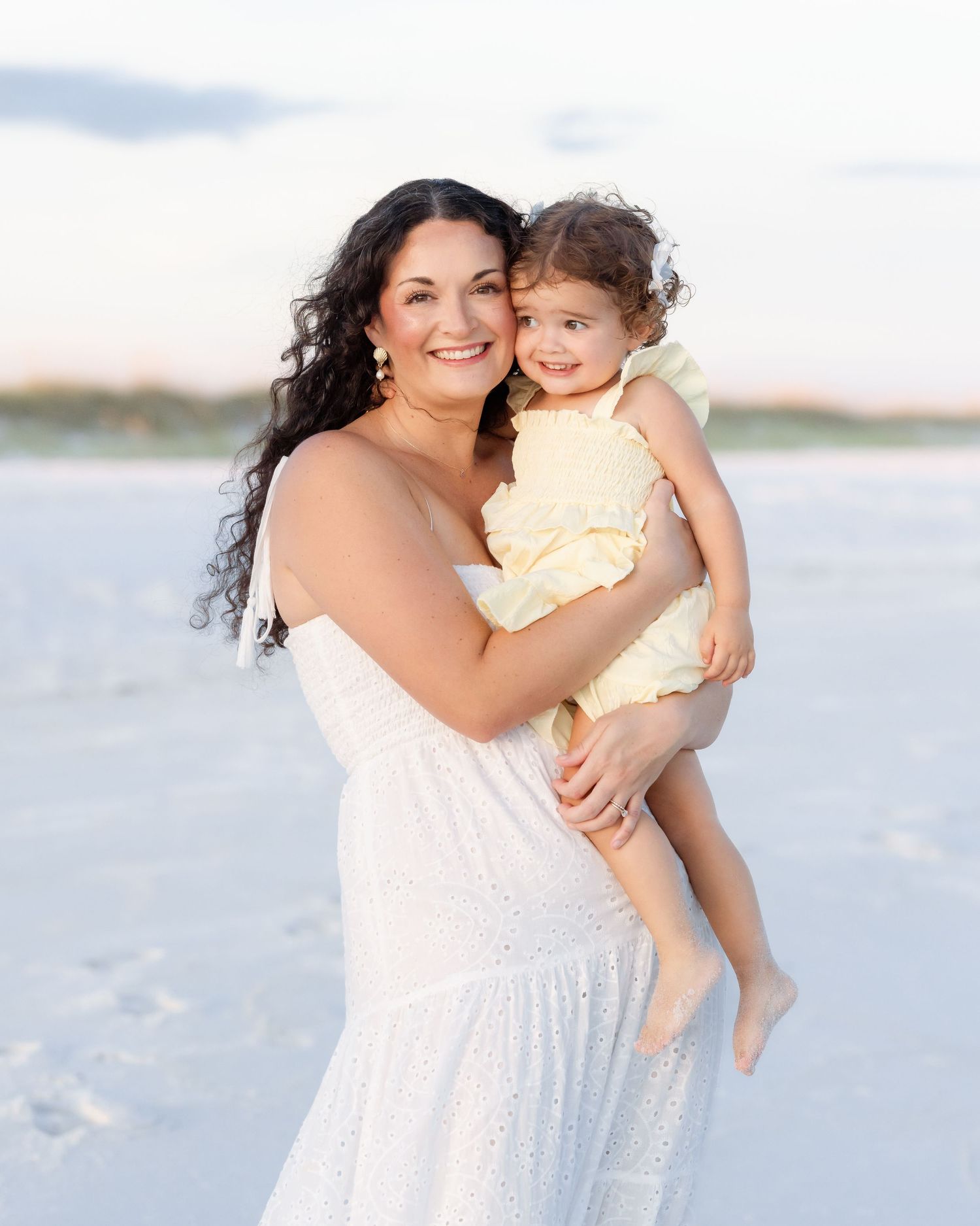 Smiling mother in white dress holding young daughter in yellow outfit on sunny beach.