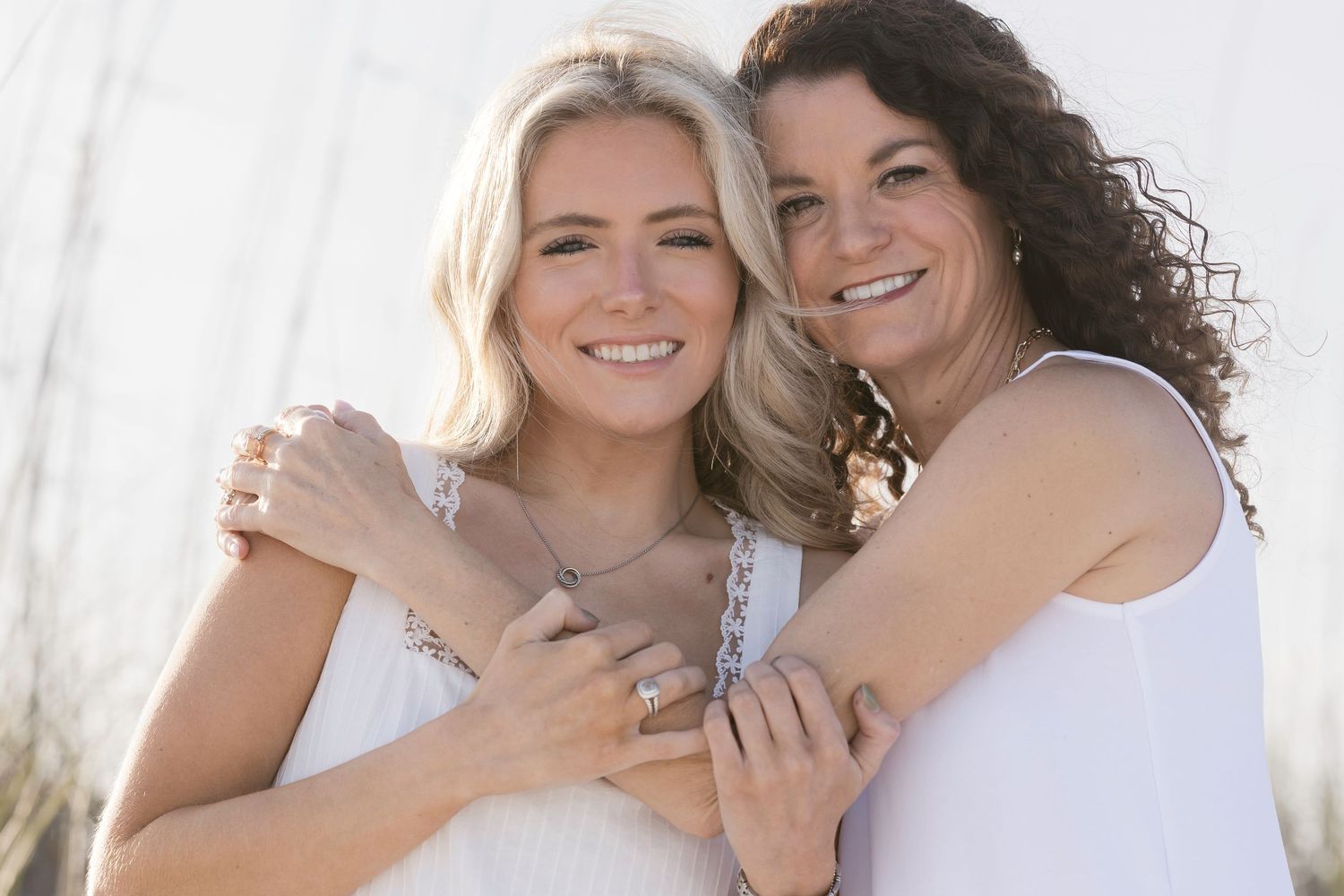 Two smiling women in white tops embrace outdoors, wearing jewelry, with a bright natural background.