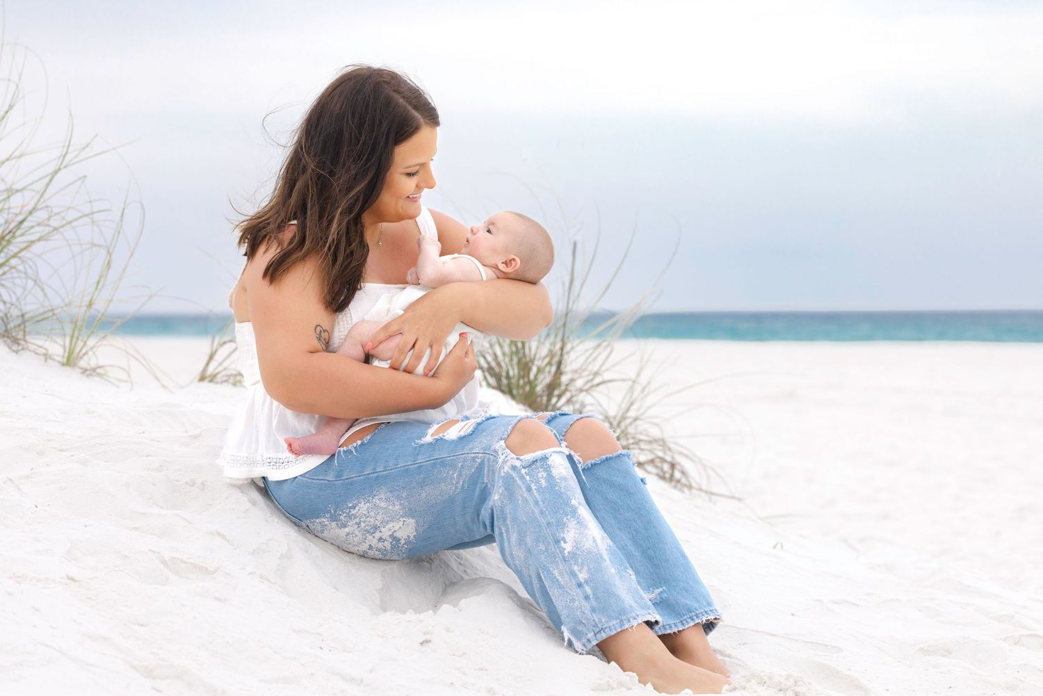 Mother breastfeeding newborn baby on white sand beach, wearing ripped jeans and white top, serene coastal setting.