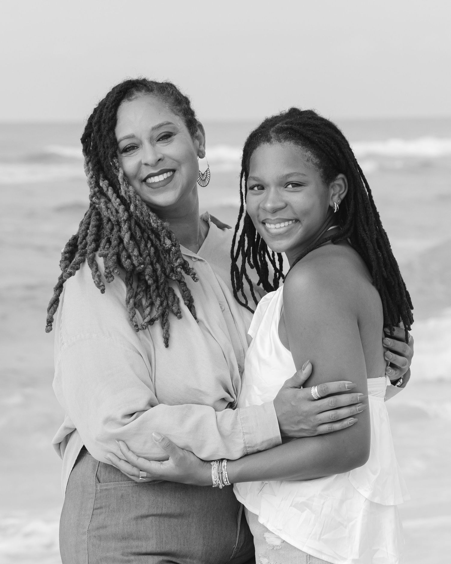 Black and white portrait of a mother and daughter with braids embracing on a beach.