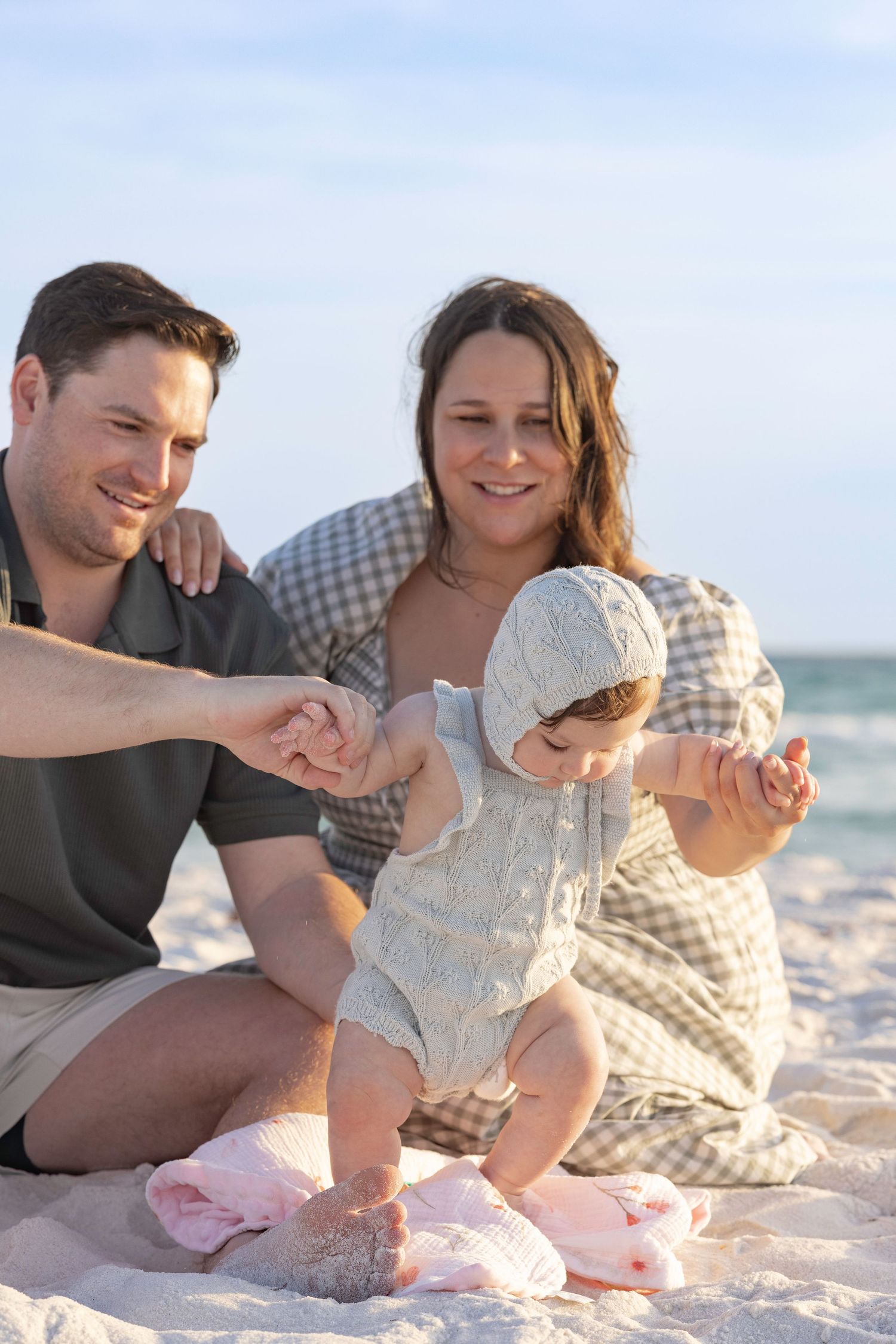 A happy young family with a baby girl sitting on a sandy beach, with the ocean in the background on a sunny day.