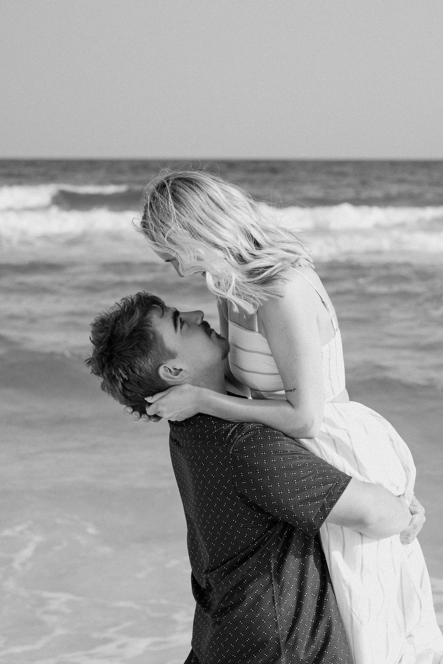 Black and white photo of a couple embracing on a beach, man lifting woman as waves crash behind them.