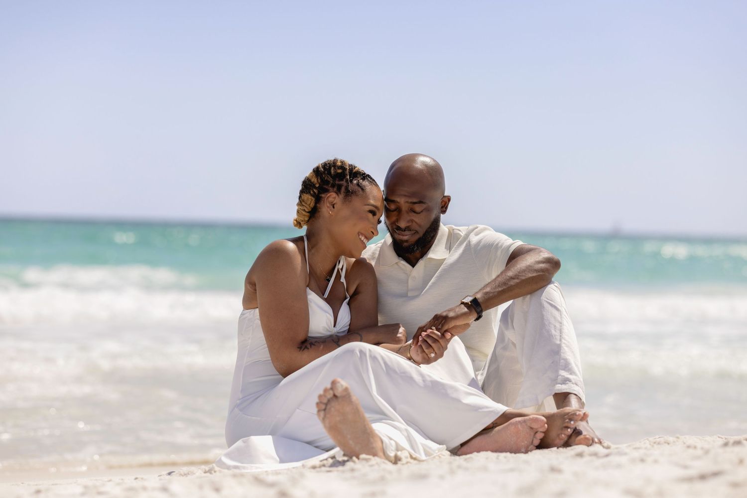 Romantic couple in white outfits sitting together on a sandy beach with turquoise ocean waves in the background.