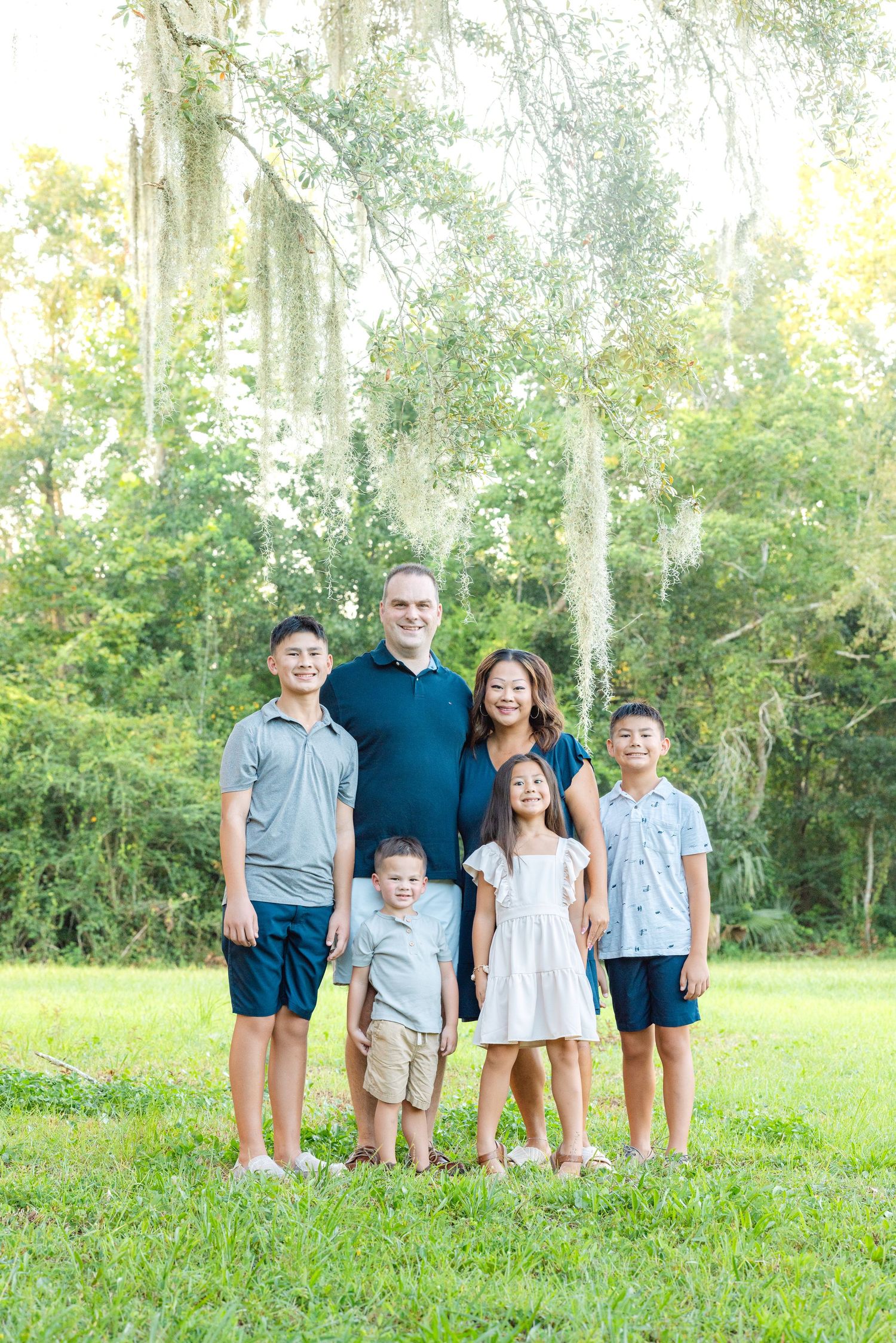Family of six posing outdoors on green grass with Spanish moss trees, featuring parents and four children in coordinated outfits.