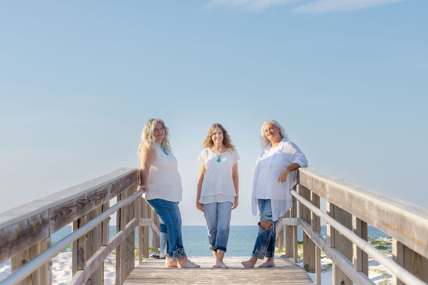 Three people stand on a wooden boardwalk along a sunny beach, framed by blue skies and white sand.