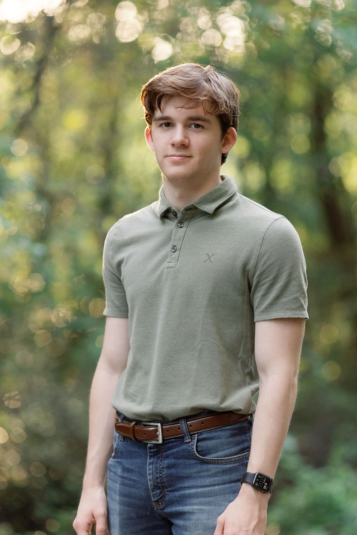 Young person in grey polo shirt and jeans standing outdoors with blurred forest bokeh background.