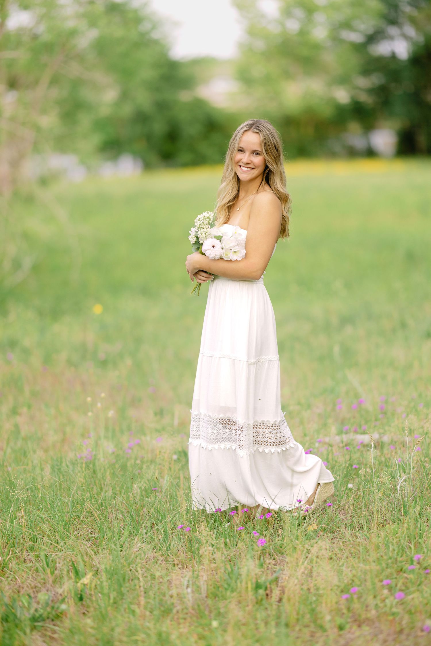 Someone wearing a white lace maxi dress poses in a sunlit meadow holding a delicate bouquet of flowers.
