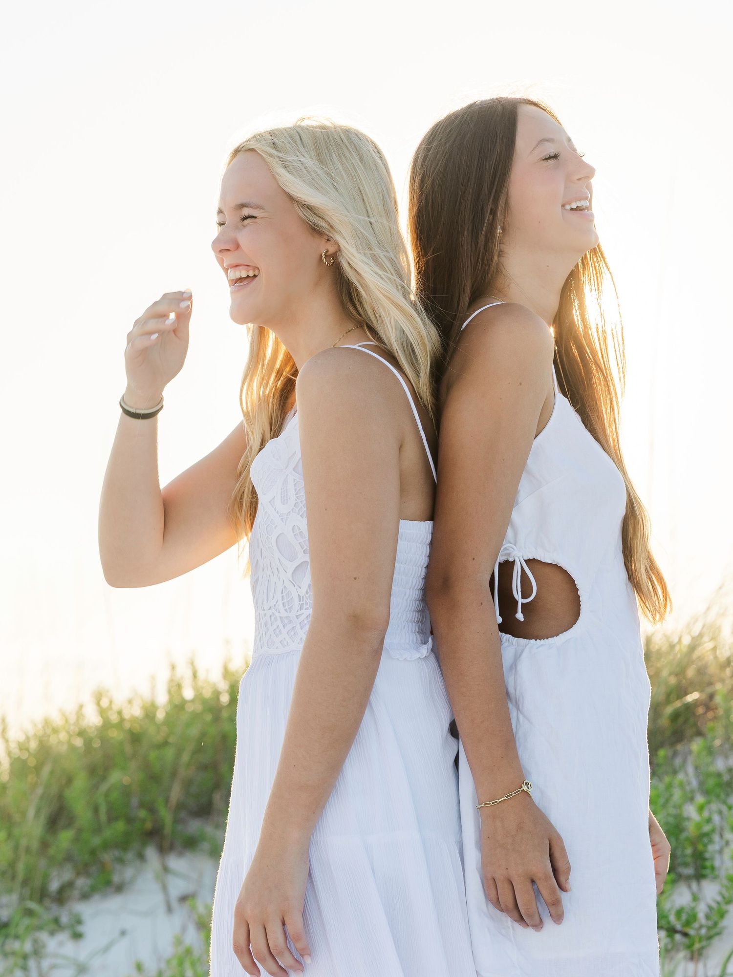 Two friends in white dresses share a joyful moment back-to-back on a sunny beach at sunset.