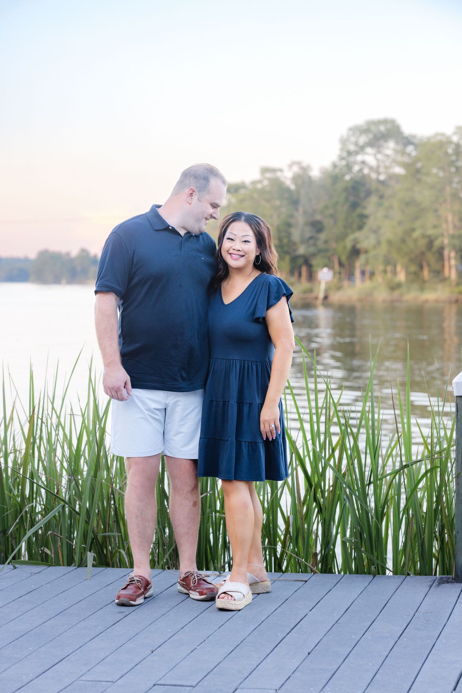 Couples standing on wooden dock by lake with tall reeds at sunset.