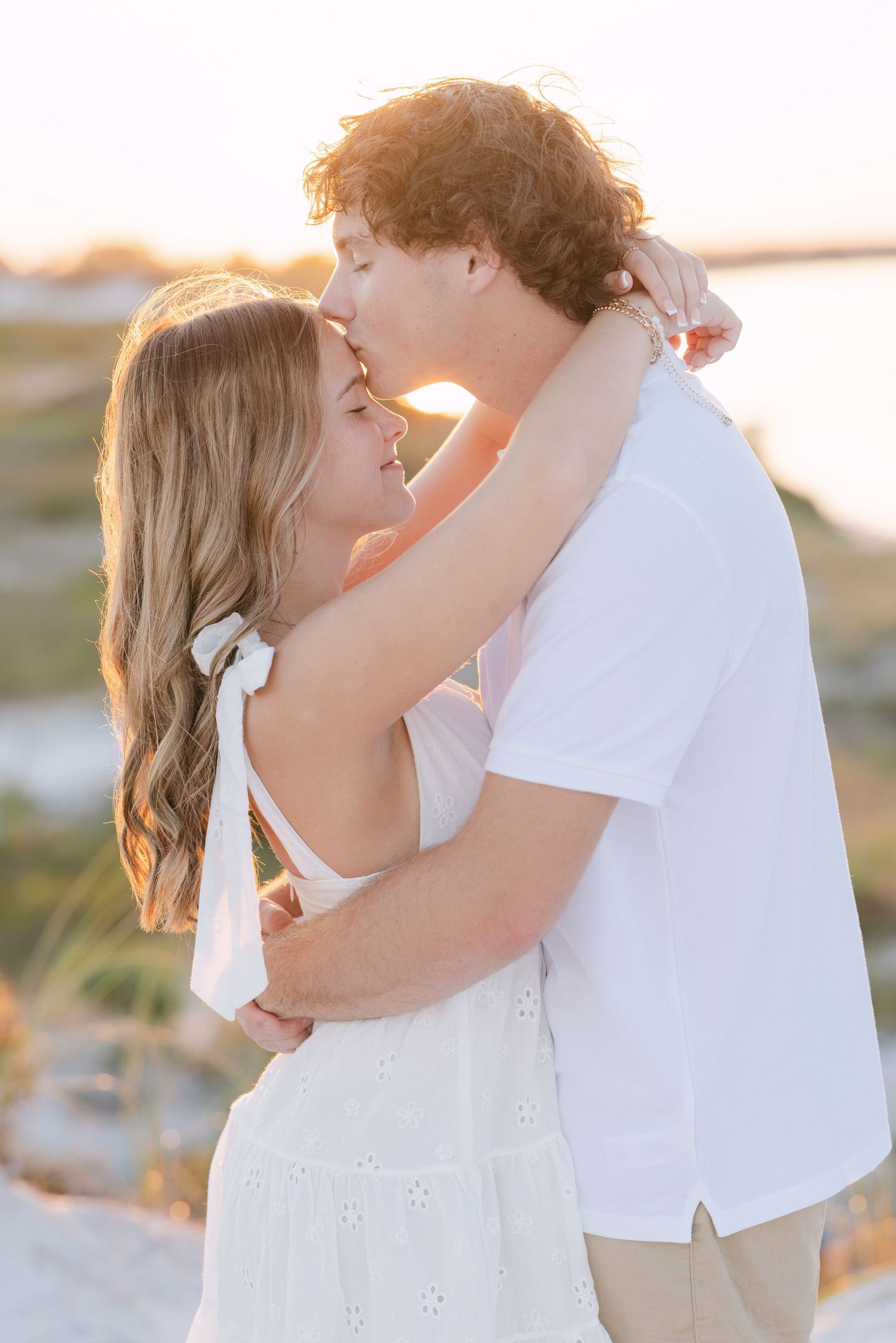 Romantic couple embracing at sunset with golden light on a beach, wearing white summer attire.