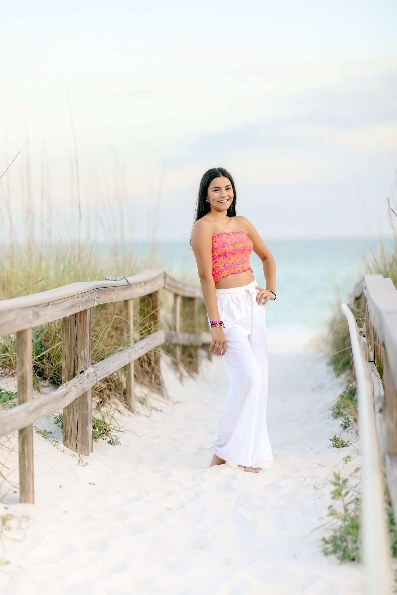 Seaside photoshoot on wooden boardwalk with white pants and coral top among beach grass and dunes.