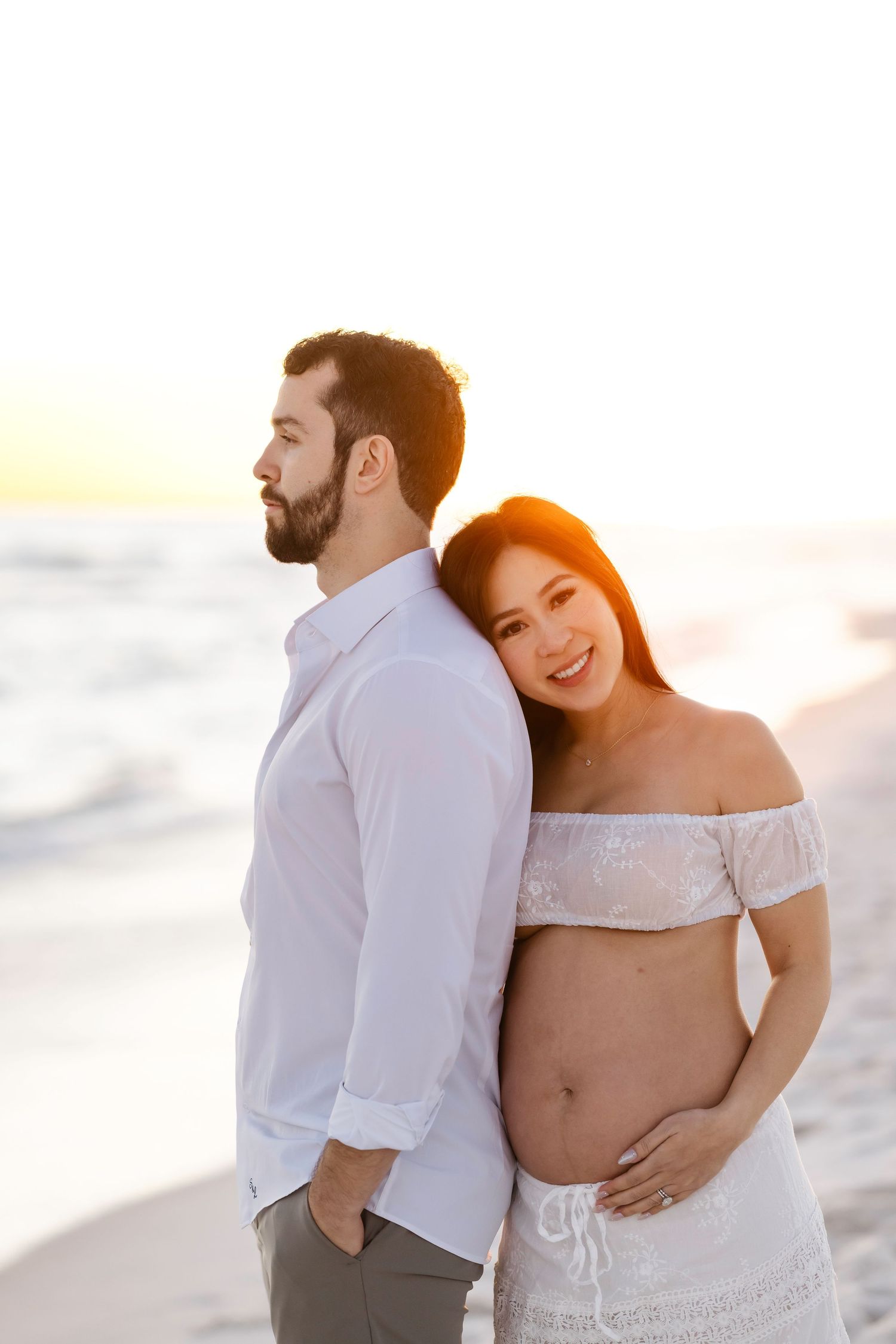 A romantic maternity photoshoot on the beach at sunset with a couple in white clothing.