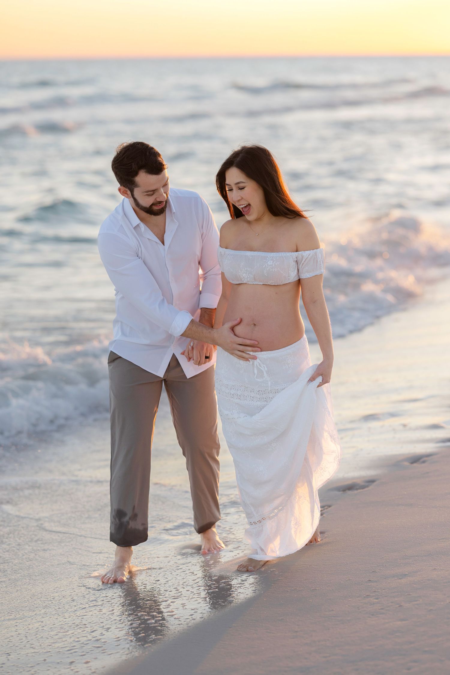 Couple wading in ocean waves at sunset wearing white and pink outfits on beach.