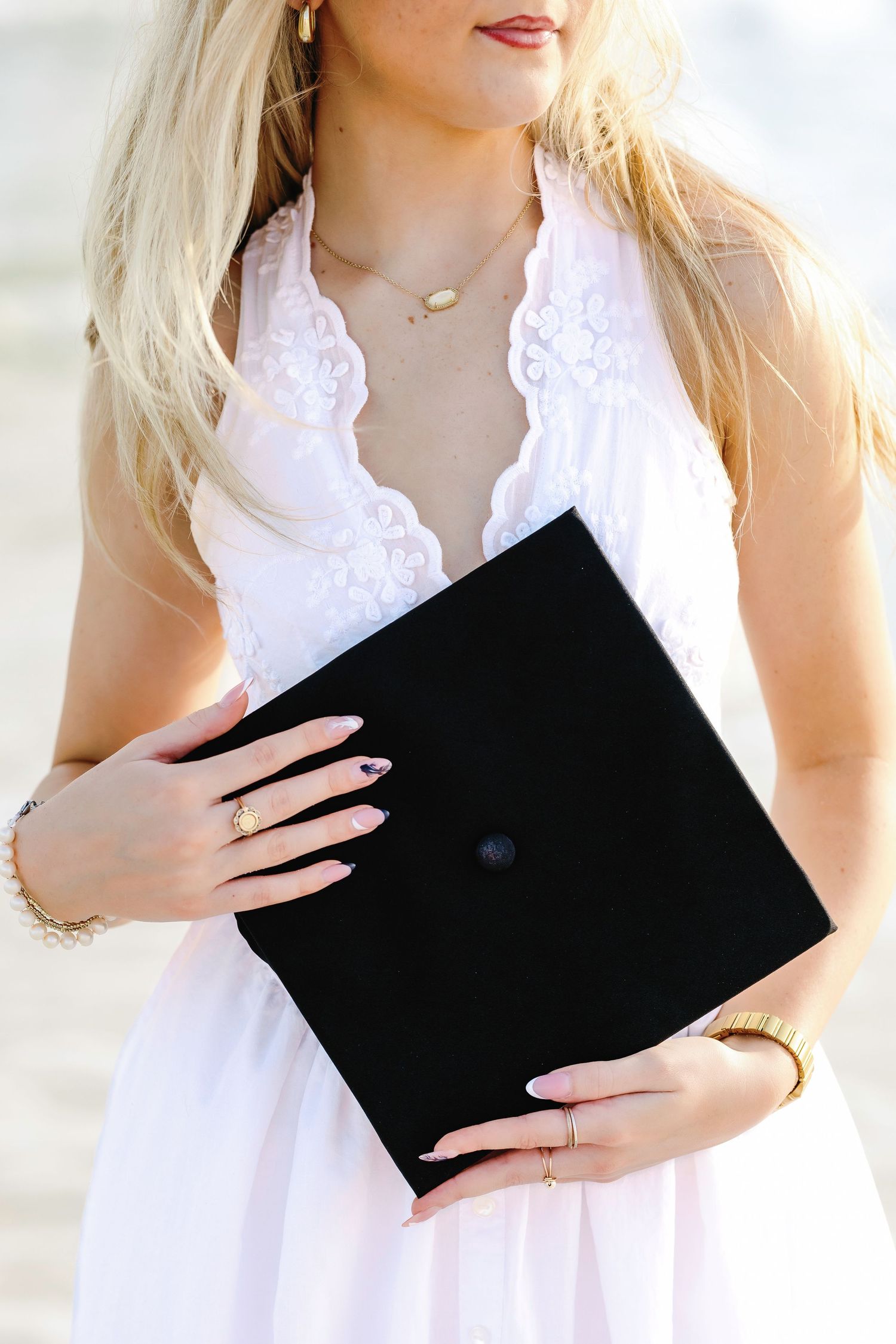 Blonde woman in white lace halter dress holding a black graduation cap, wearing gold jewelry.
