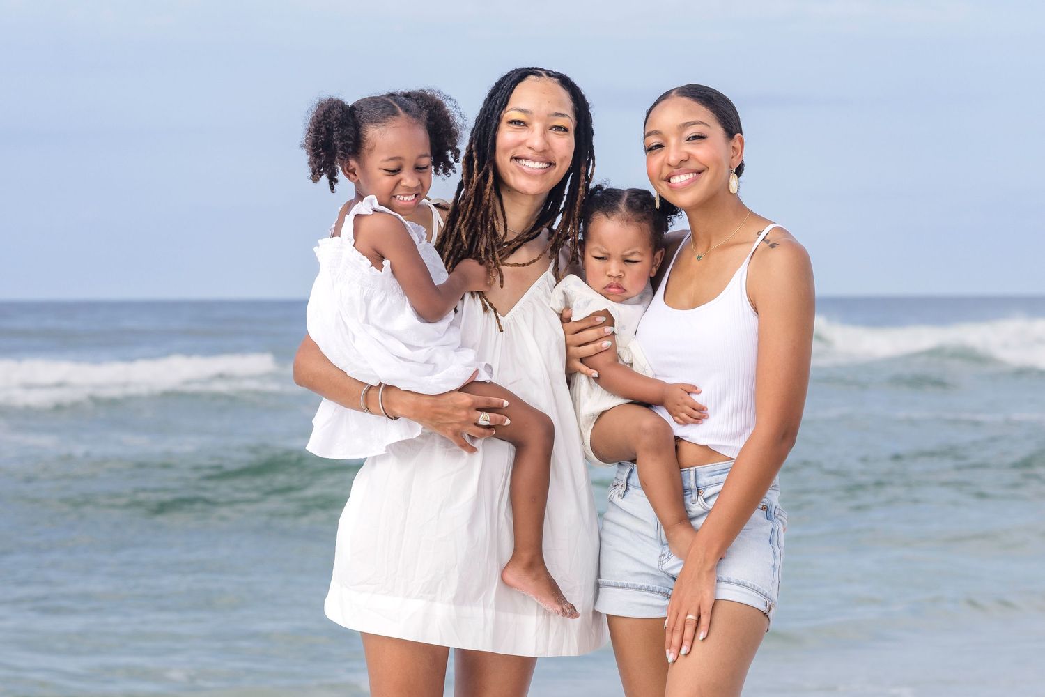 Two women and two toddlers smile together on a sunny beach with ocean waves in the background.