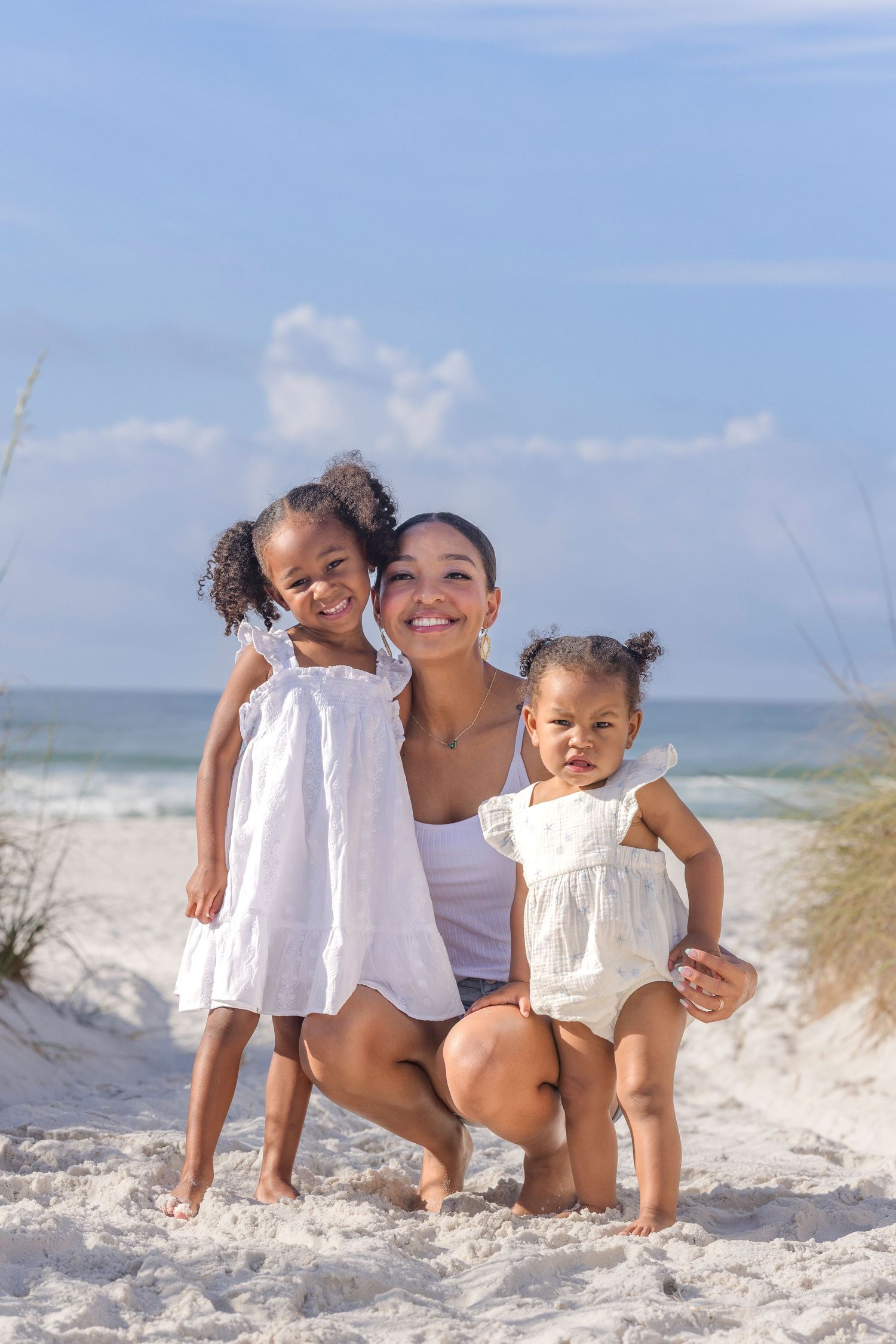 A smiling mother crouches on a white sandy beach with her two young daughters in matching white dresses on a sunny day.
