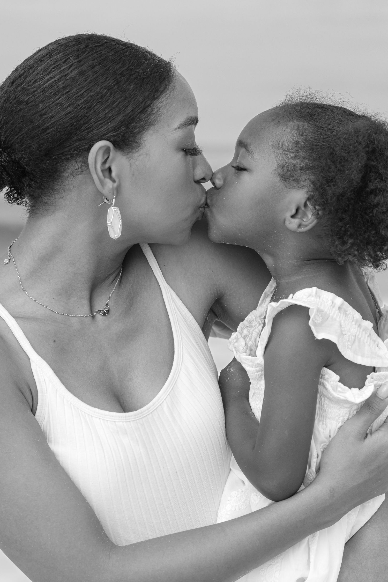 Black and white photo of a mother kissing her young daughter, both wearing white, sharing a tender moment.